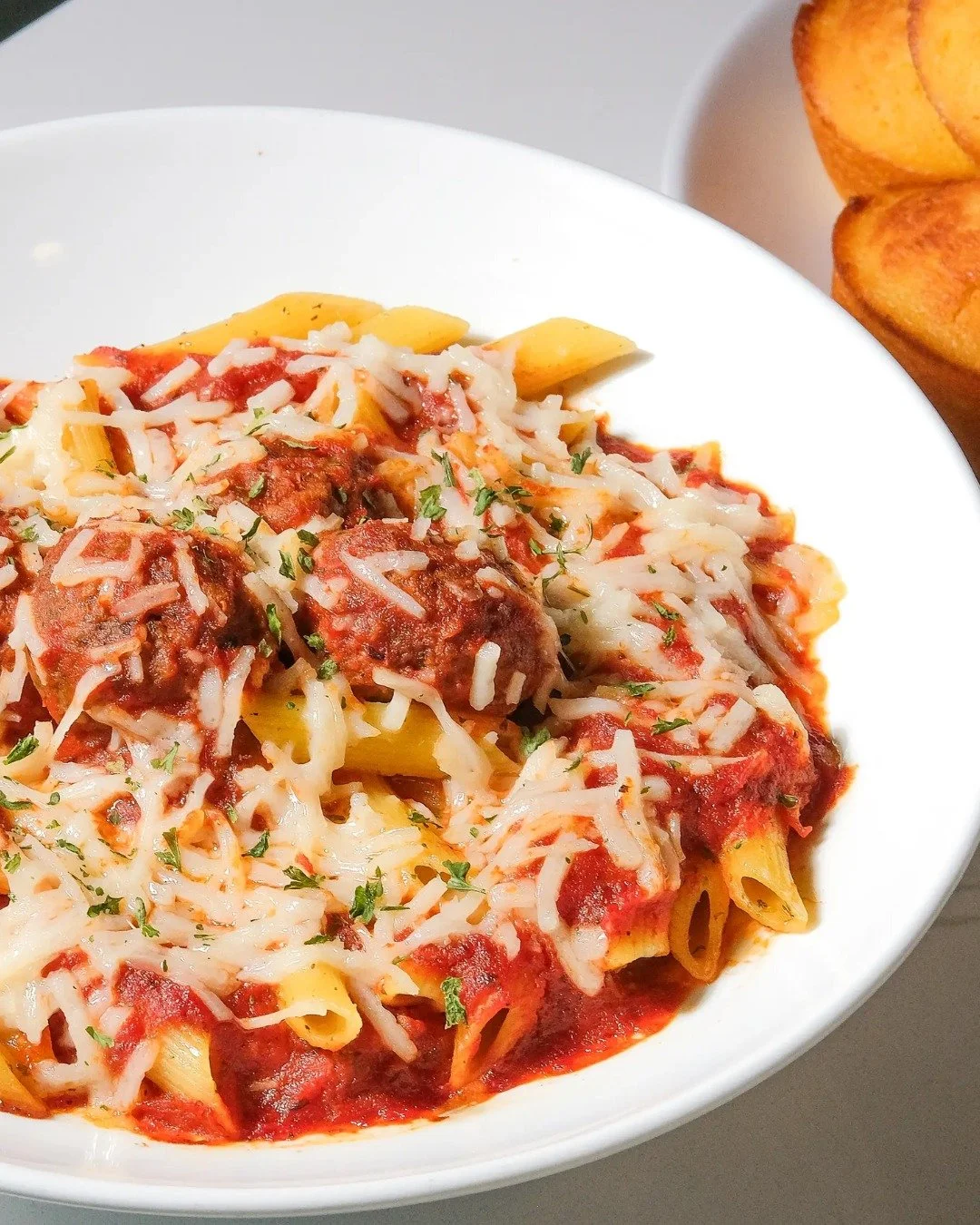Bowl of baked pasta with meatballs, tomato sauce, melted cheese, and chopped herbs, served with baked bread on the side.