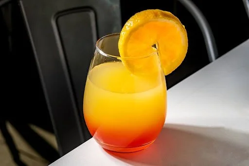 A tropical drink in a clear glass with a slice of orange on the rim, featuring a gradient of red, orange, and yellow colors.