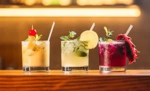 Three colorful cocktails on a bar countertop with straws and garnishes, illuminated by warm lighting.