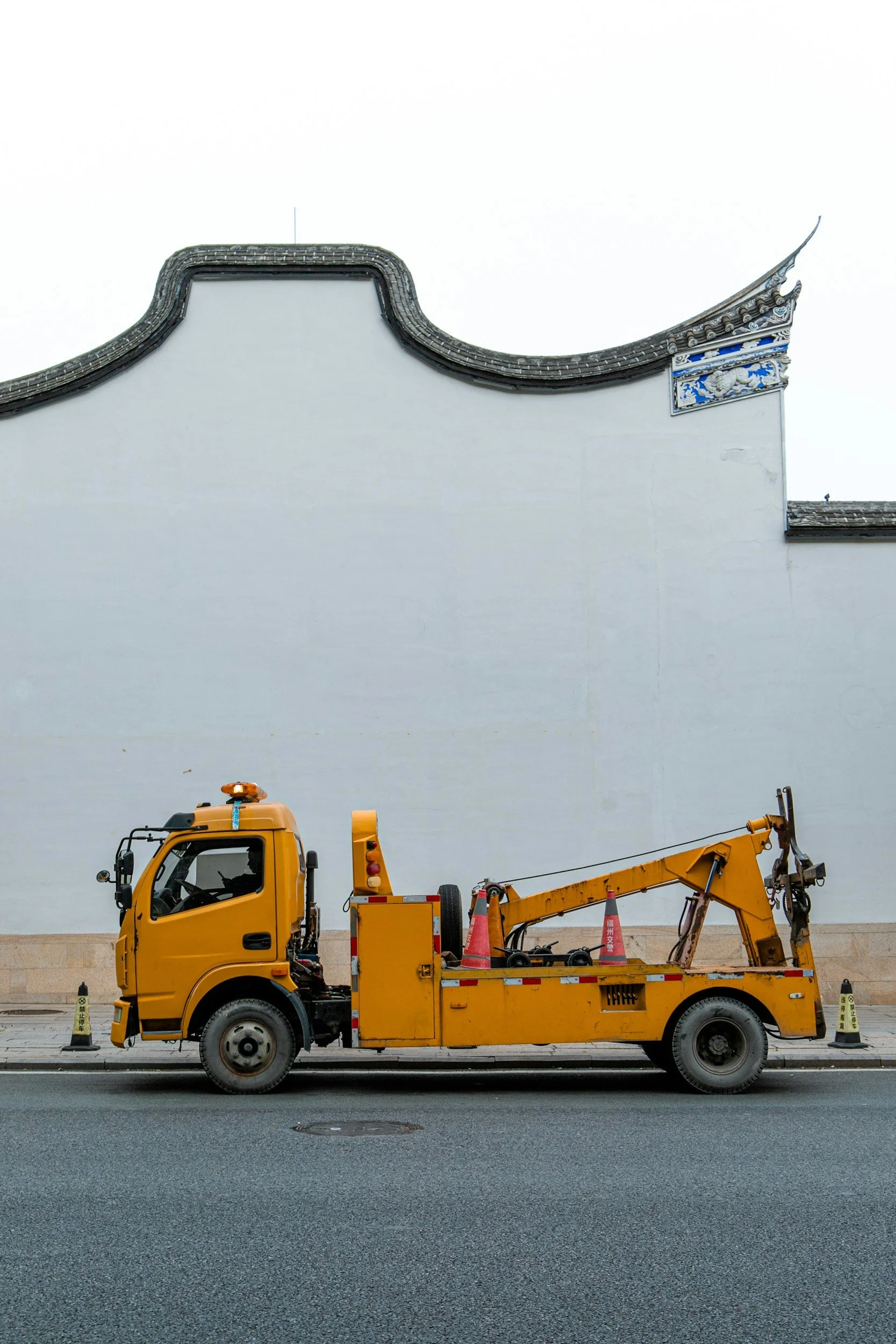 Yellow tow truck parked on street in front of a large white wall with traditional Asian architectural features.