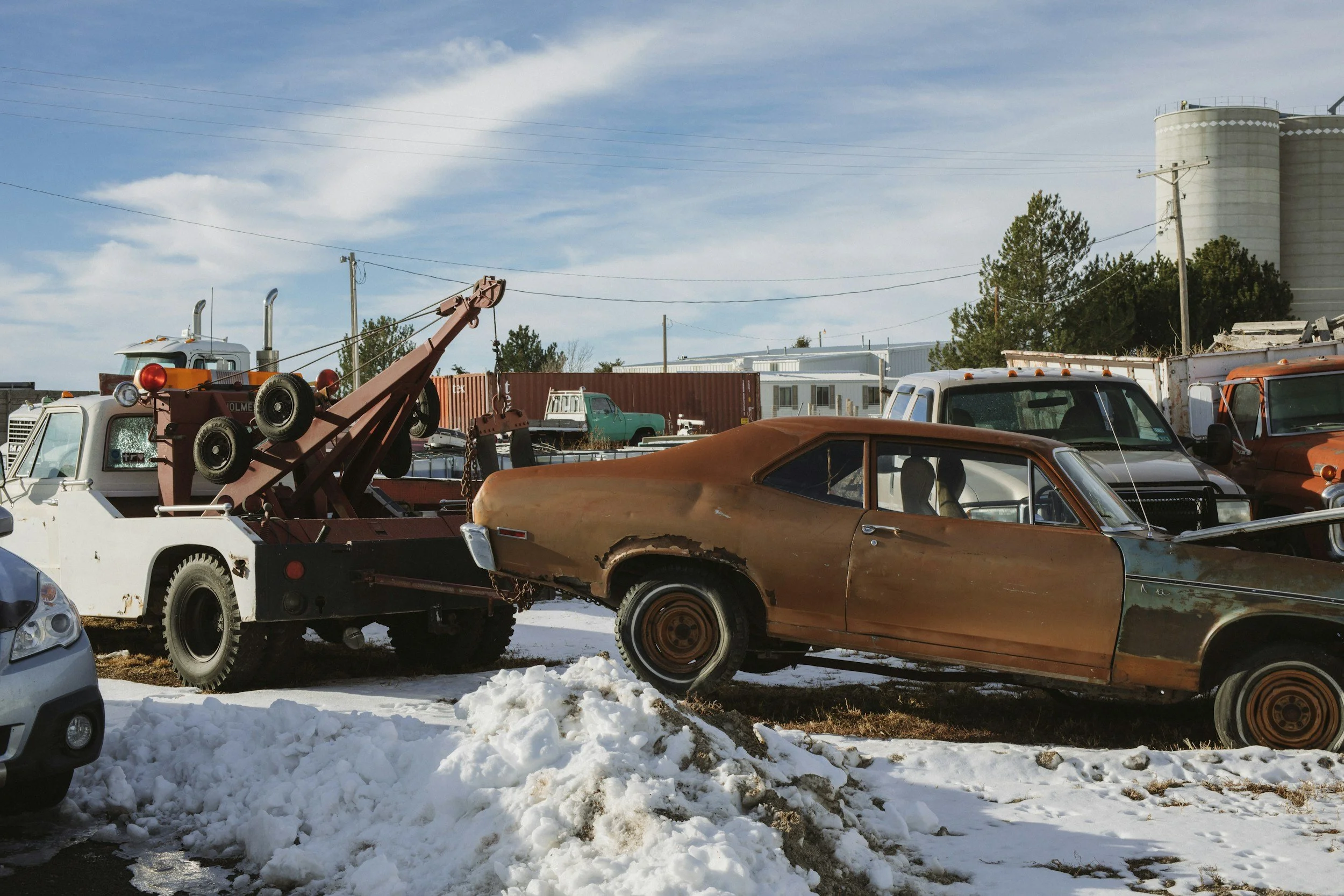 Old rusty brown car parked next to a tow truck in a junkyard with snow on the ground, other vintage vehicles and industrial buildings in the background.