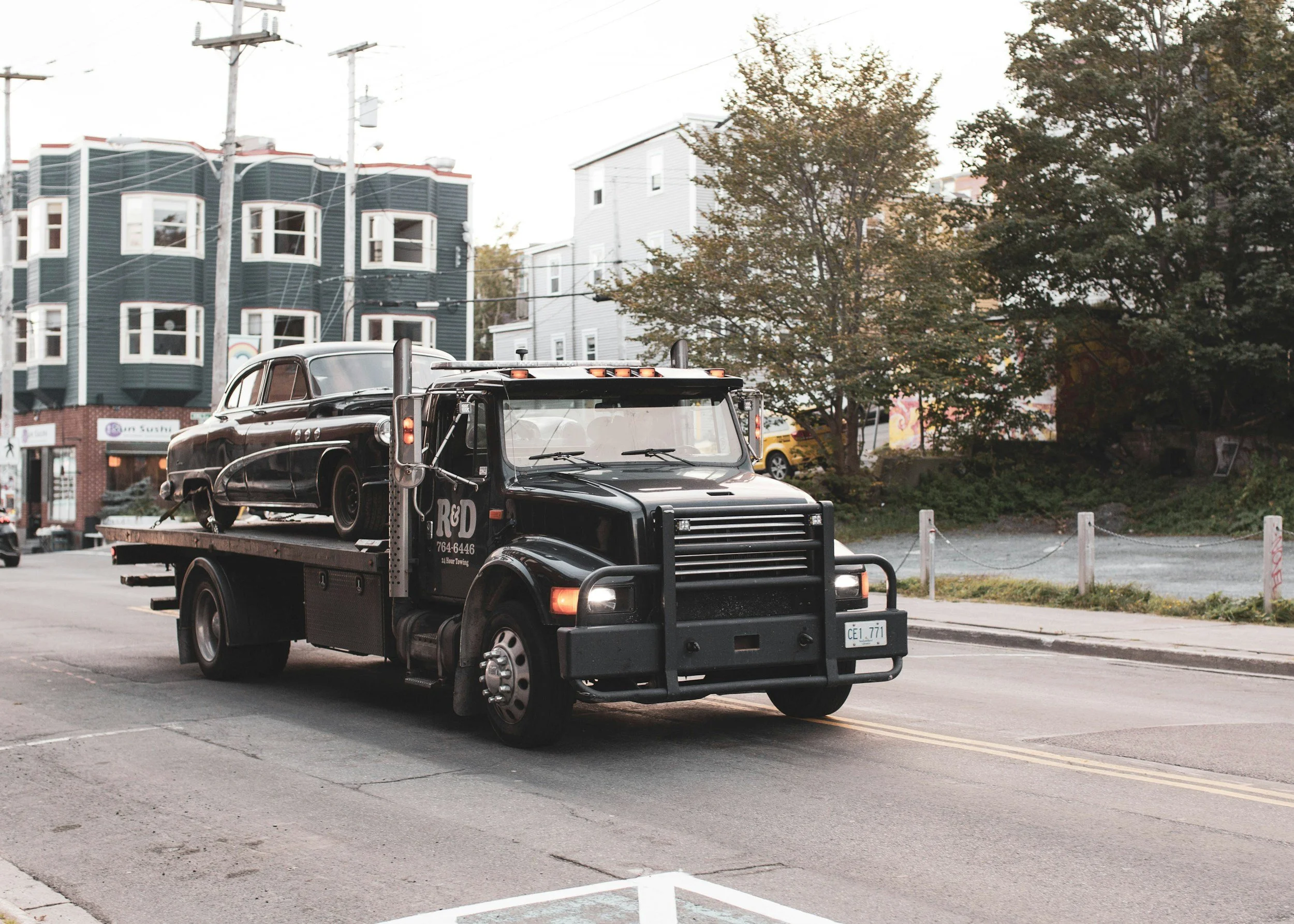 Black tow truck transporting a vintage black car on a city street with trees and buildings in the background.