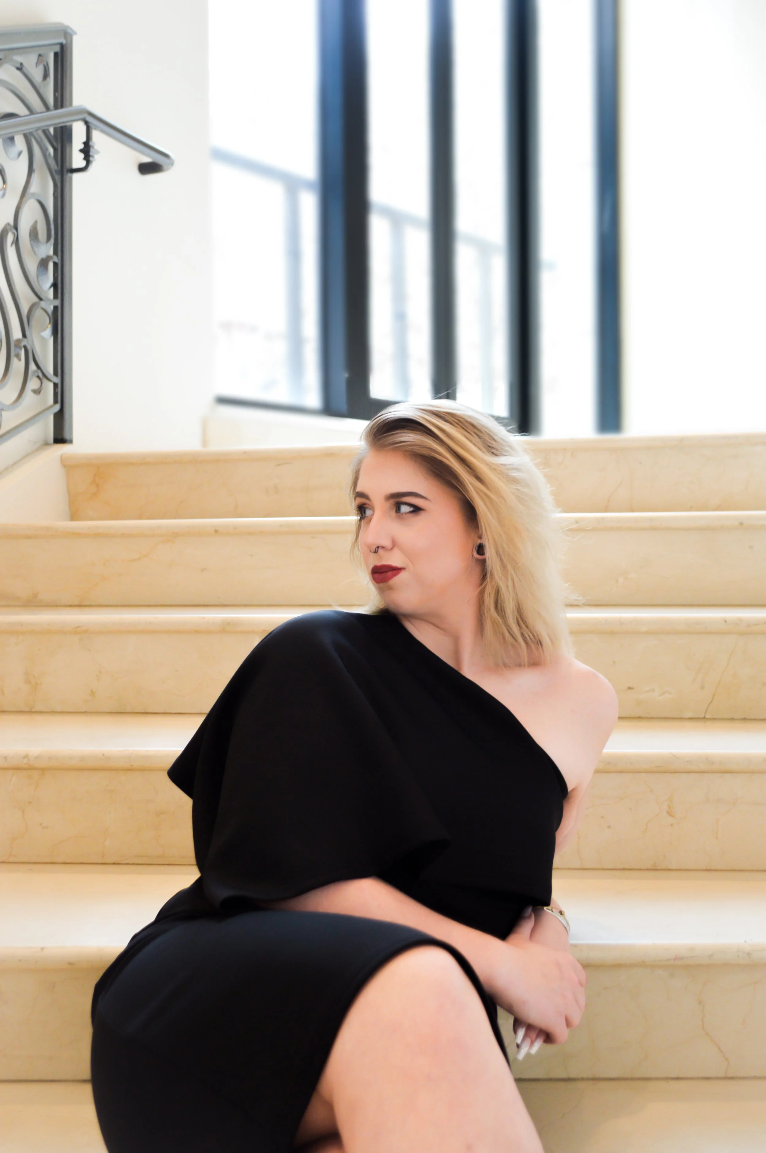 A woman with blonde hair and red lipstick sitting on marble stairs with a black off-shoulder dress. She has a septum piercing and earrings, looking to her left with a neutral expression. In the background, there are large windows and a decorative iron railing.