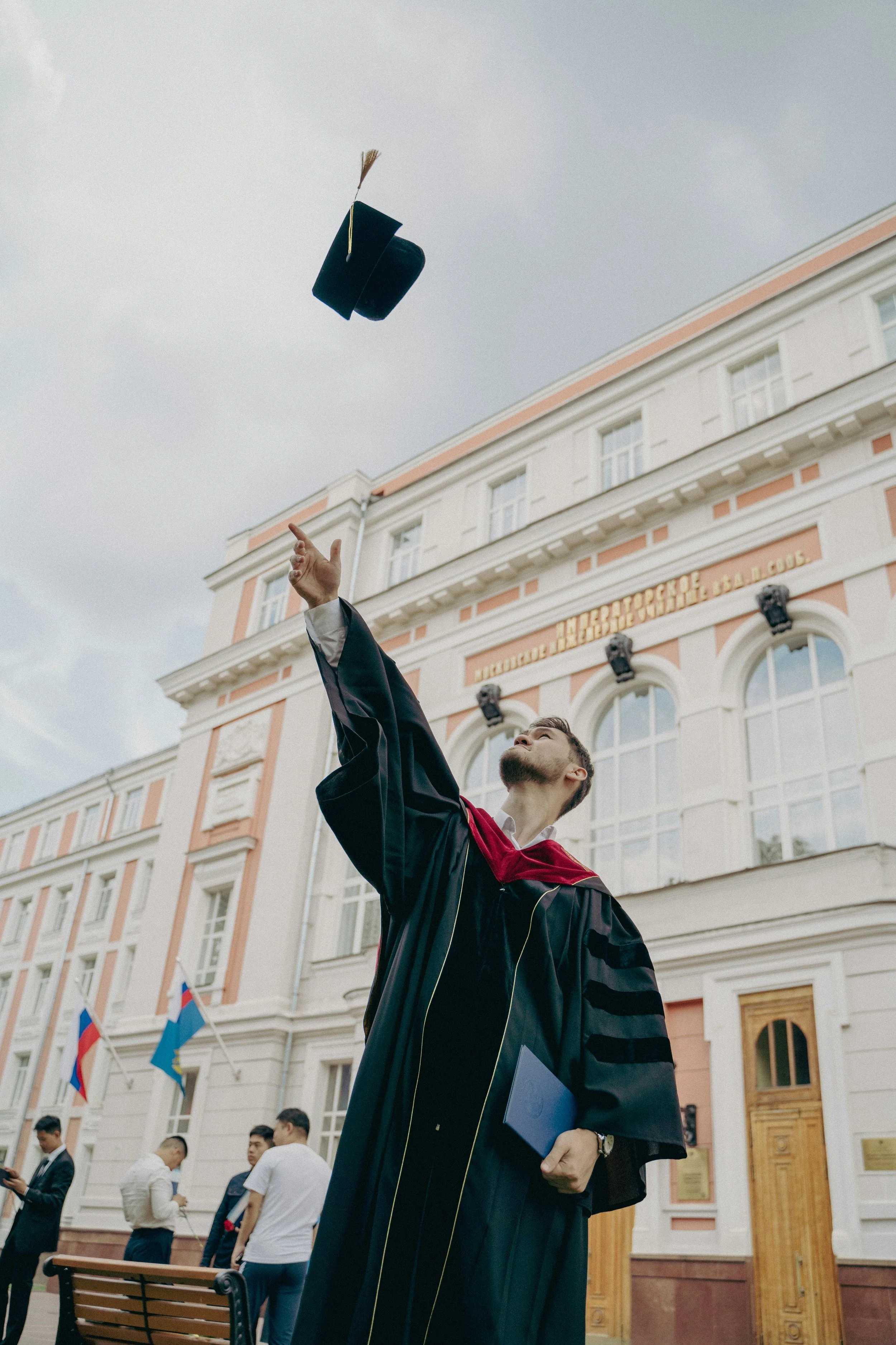 A graduate in a black gown and red hood tosses his cap in the air during an outdoor graduation ceremony, with a historic building and flags in the background.