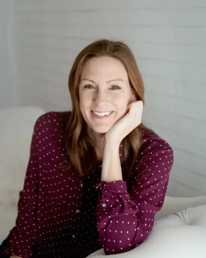 A smiling woman with reddish hair, wearing a maroon polka-dot shirt, resting her chin on her hand, sitting on a bed in a softly lit room with white wooden walls.