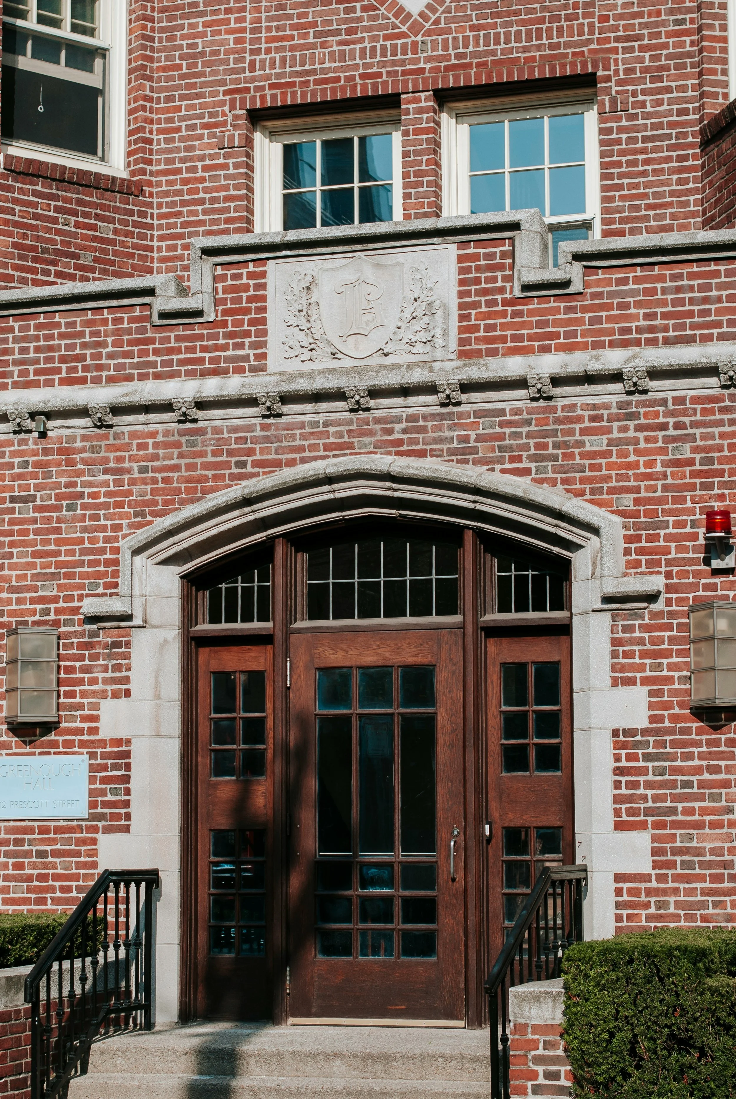 Brick building with a wooden door, arched window, and stone decorative elements. Two small windows above the door, a stone crest with shield and leaf designs, and exterior lights on either side of the door.