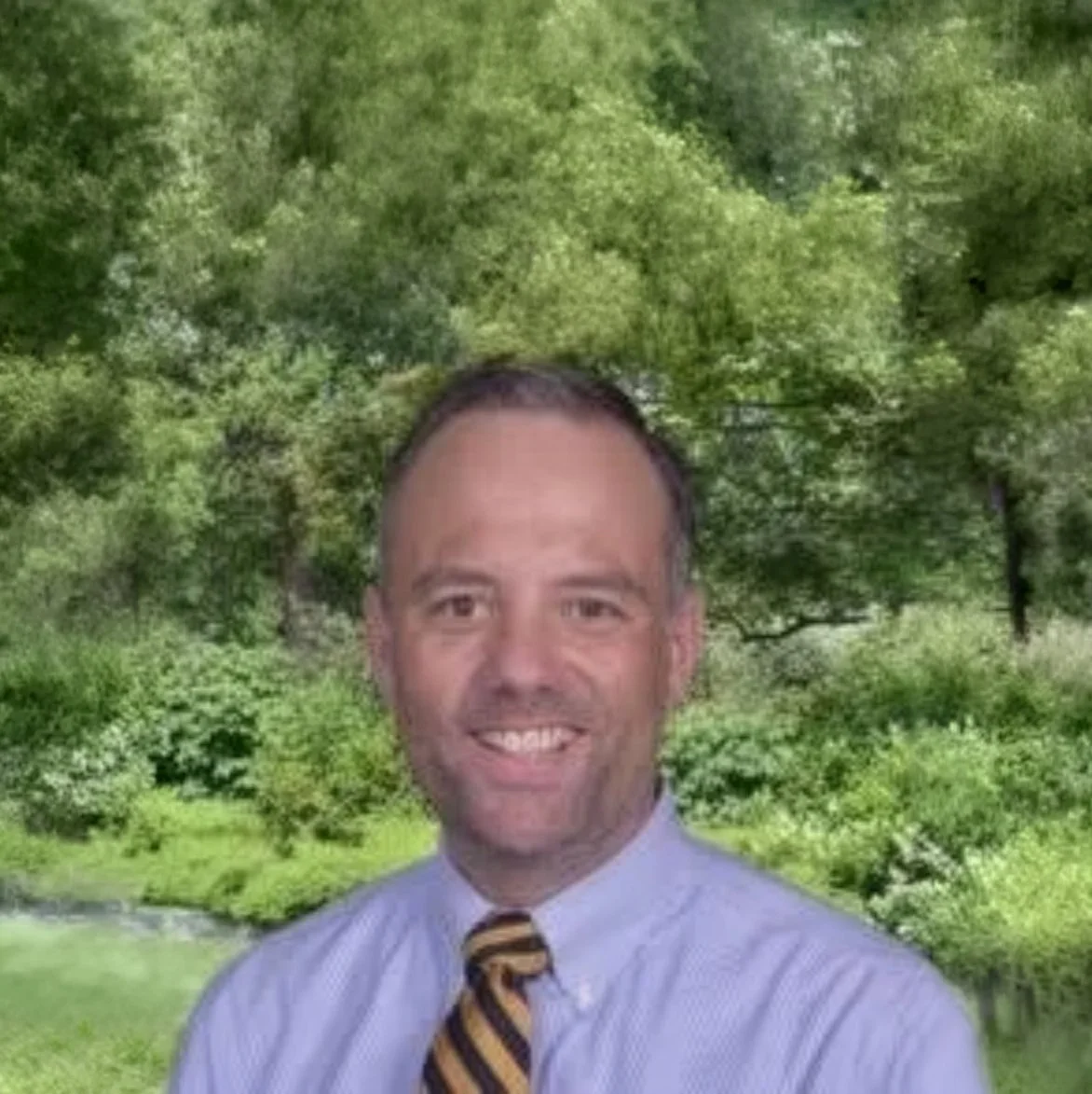 A smiling man with short dark hair, wearing a light blue dress shirt and a striped tie, standing outdoors with a background of green trees and foliage.