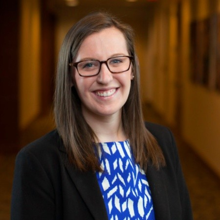 A smiling woman with glasses and long brown hair, wearing a black blazer over a blue and white patterned shirt, standing in a hallway with warm lighting and bookshelves.