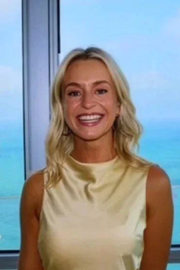 A woman with blonde hair smiling, wearing a sleeveless yellow top, standing inside with a large window behind her showing a body of water and a cloudy sky.