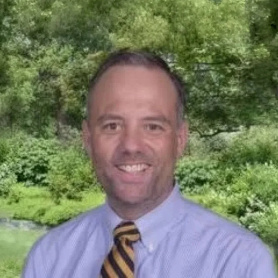 A man with short dark hair, a beard, and a broad smile, wearing a light blue dress shirt and a striped yellow and black tie, standing outdoors with a lush green background of trees and foliage.