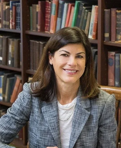 A woman with dark brown hair smiling, sitting in front of a bookshelf filled with books.