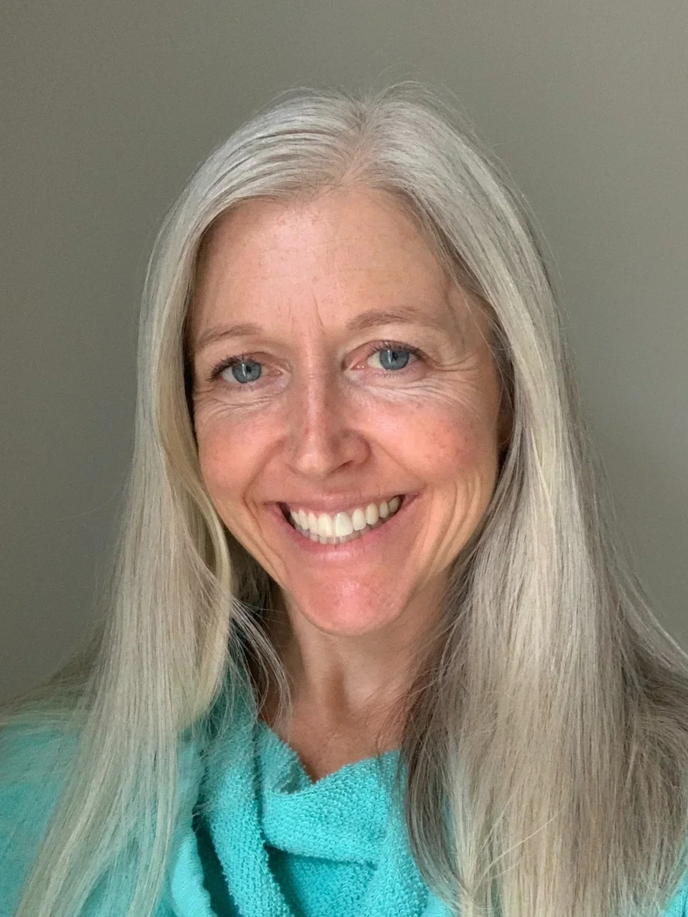 A smiling woman with long gray hair and blue eyes wearing a light teal top.