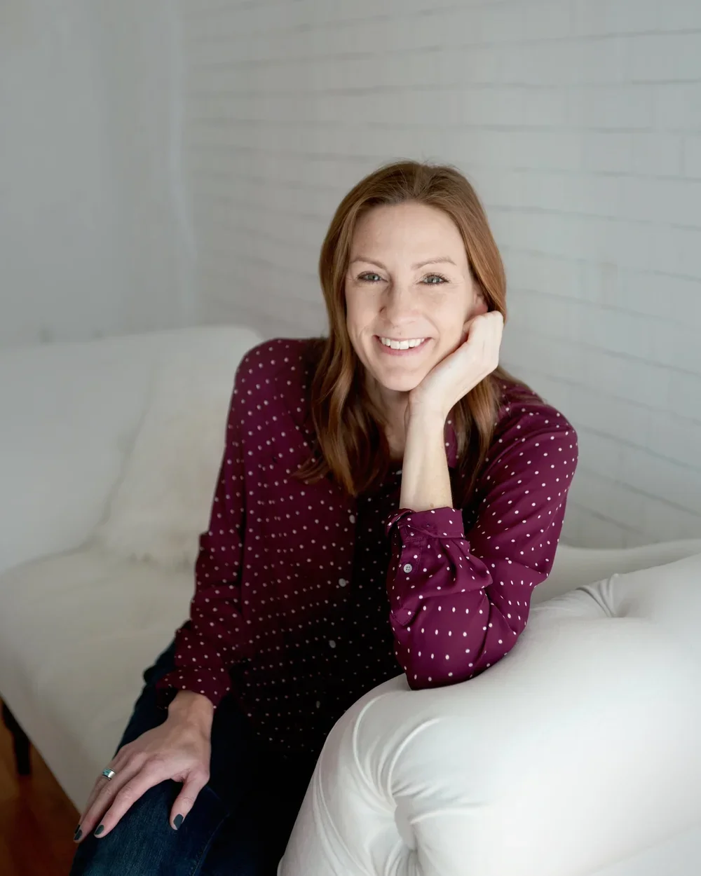A woman with shoulder-length reddish-brown hair, wearing a maroon blouse with white polka dots, smiling and resting her chin on her hand while sitting on a white sofa in a bright room with white brick walls.