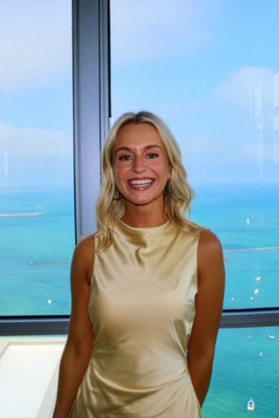 A smiling woman with blonde hair in a sleeveless light yellow top standing indoors with a large window showing a blue ocean and sky background.