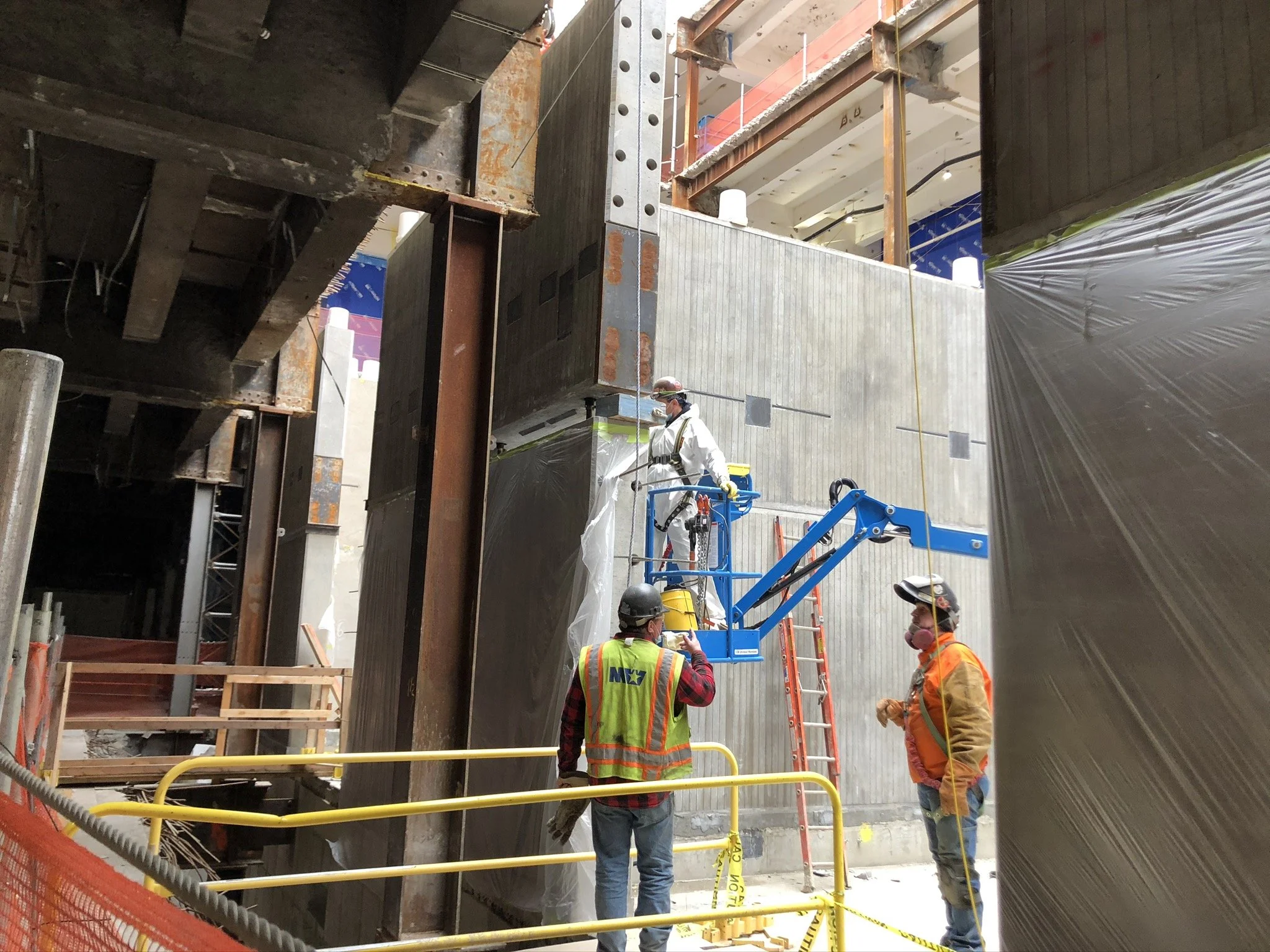 Construction workers at a building site using a blue boom lift to work on a concrete wall. Two workers stand on the ground wearing safety gear, while one worker is on the lift, working on the wall. Safety barriers and ladders are visible.