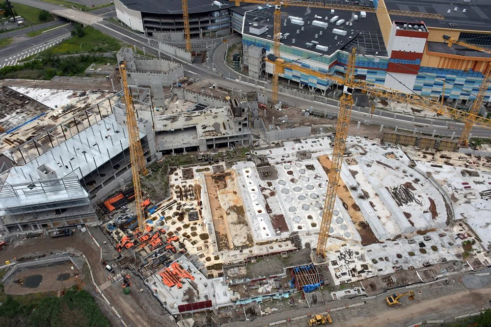Aerial view of a construction site with multiple cranes and a partially built multi-story building, with surrounding roads, some green areas, and a colorful modern building in the background.