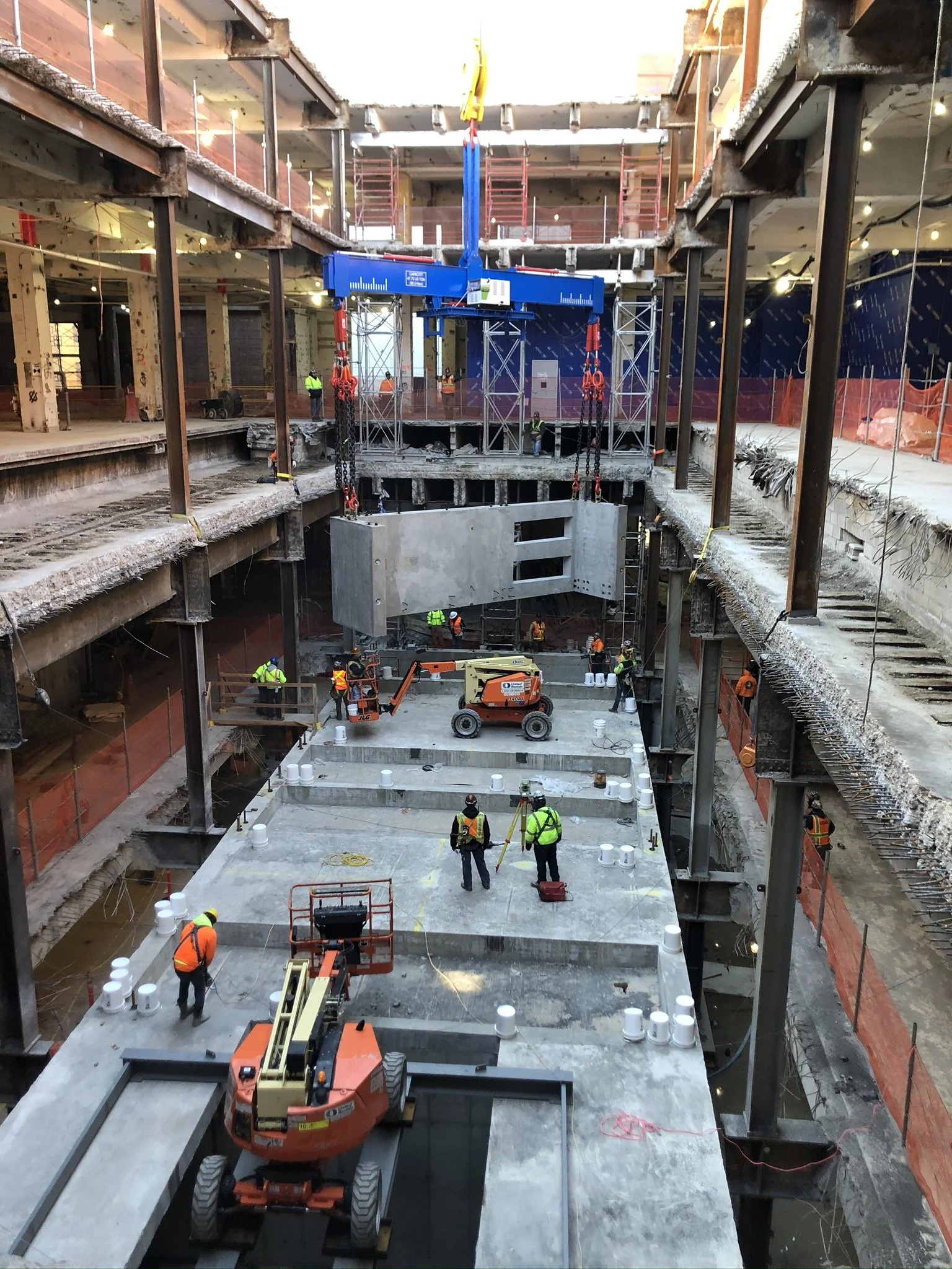 Construction workers and equipment inside a large building under construction, with a large concrete structure being lowered by a crane.