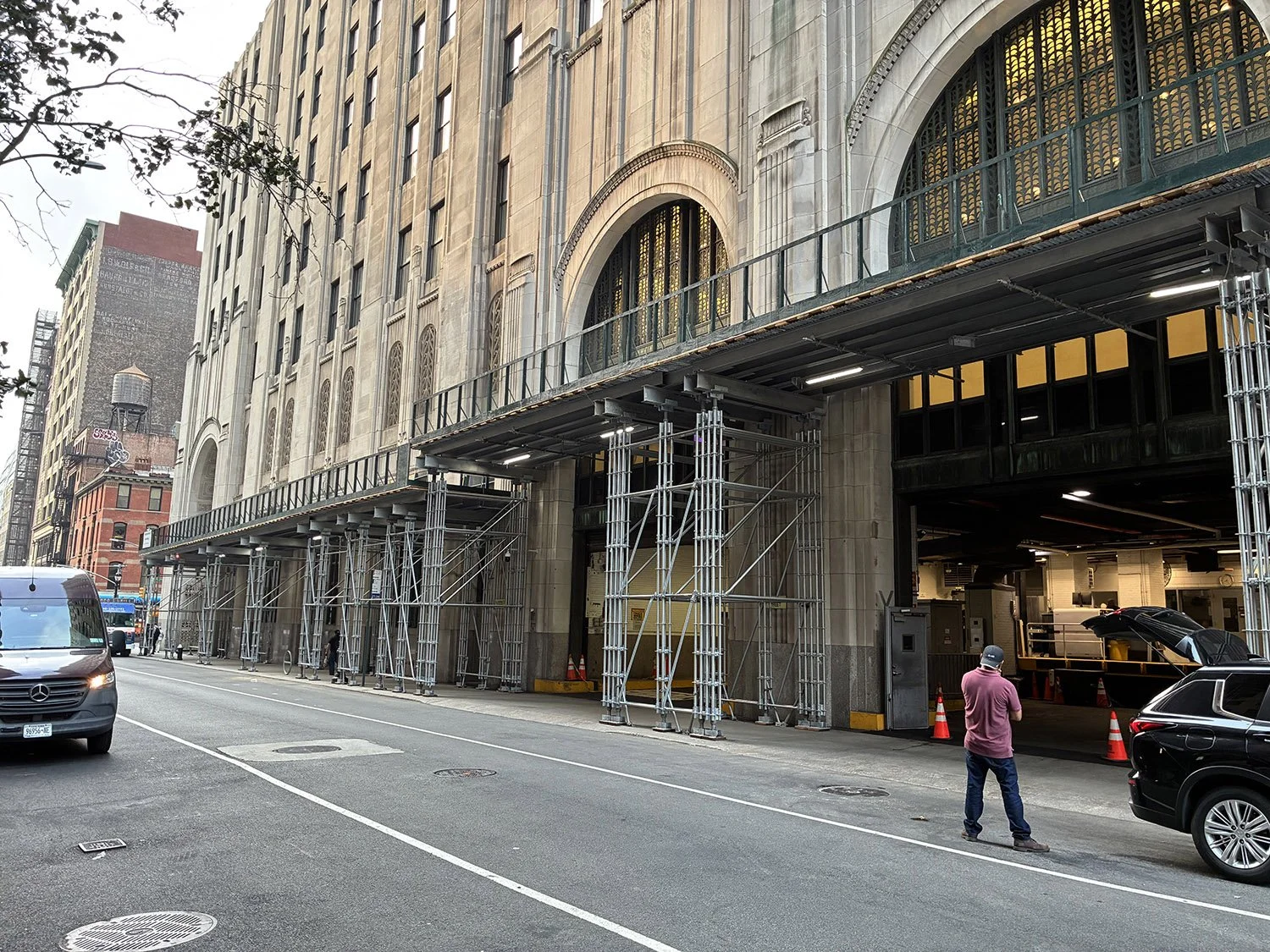 Street view of a building under construction with scaffolding along the sidewalk, cars parked on the street, and a man standing near a black vehicle.