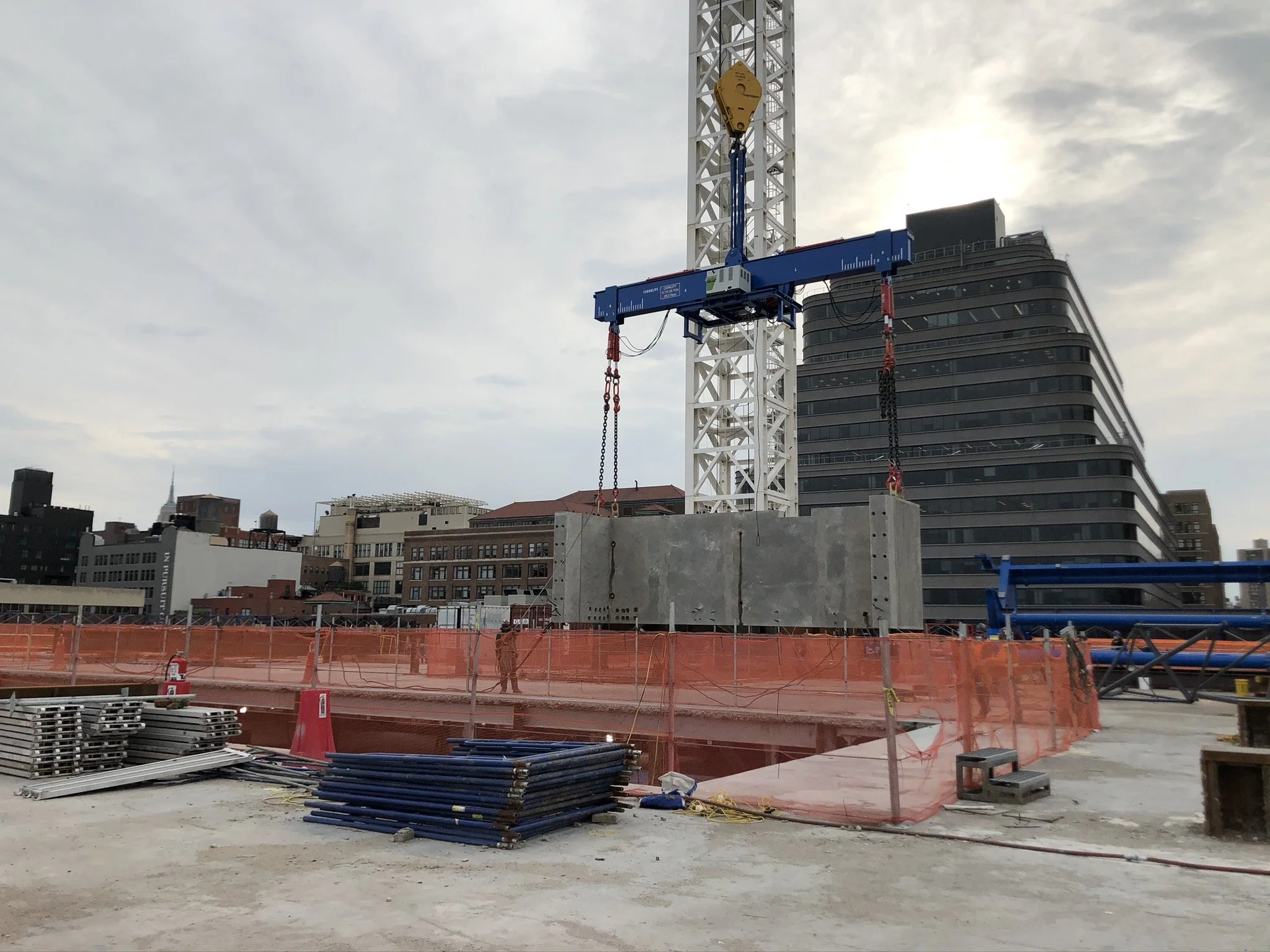 Construction site with a tower crane lifting a large concrete slab, surrounded by orange safety fencing, with city buildings and cloudy sky in the background.