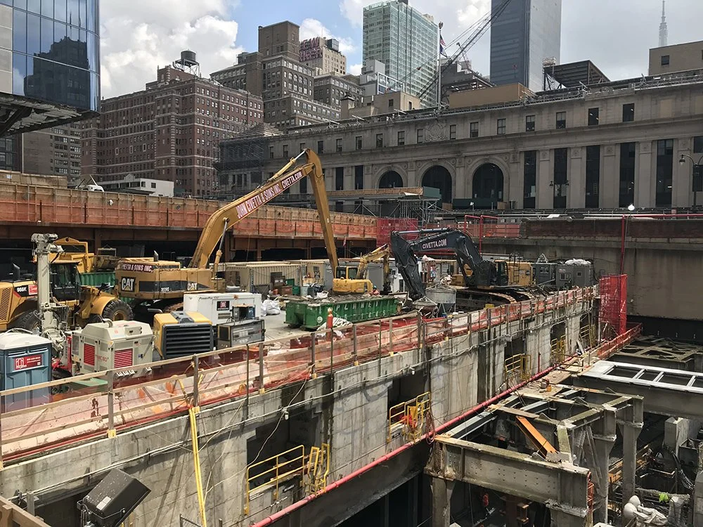 Construction site with excavators, machinery, and safety barriers in an urban cityscape with tall buildings and a cloudy sky.