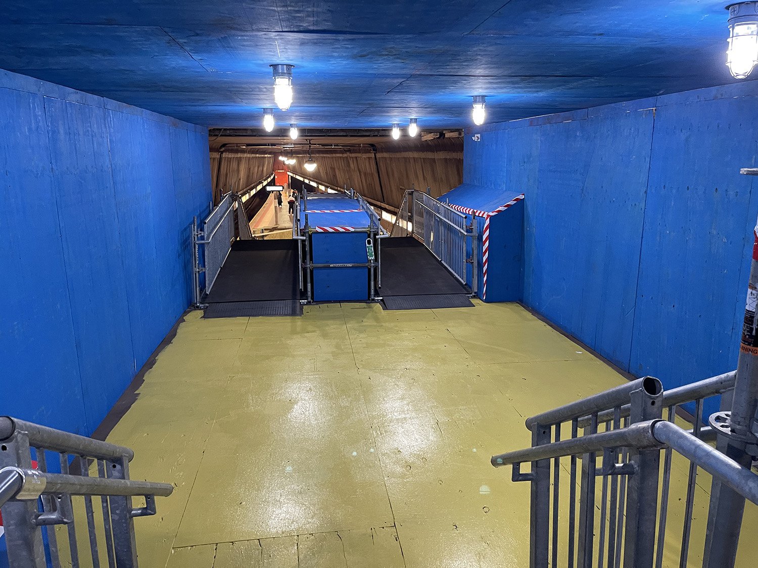 An indoor pedestrian walkway with a ramp for wheelchair access, blue walls, yellow floor, and overhead lighting, leading to an underground station platform.