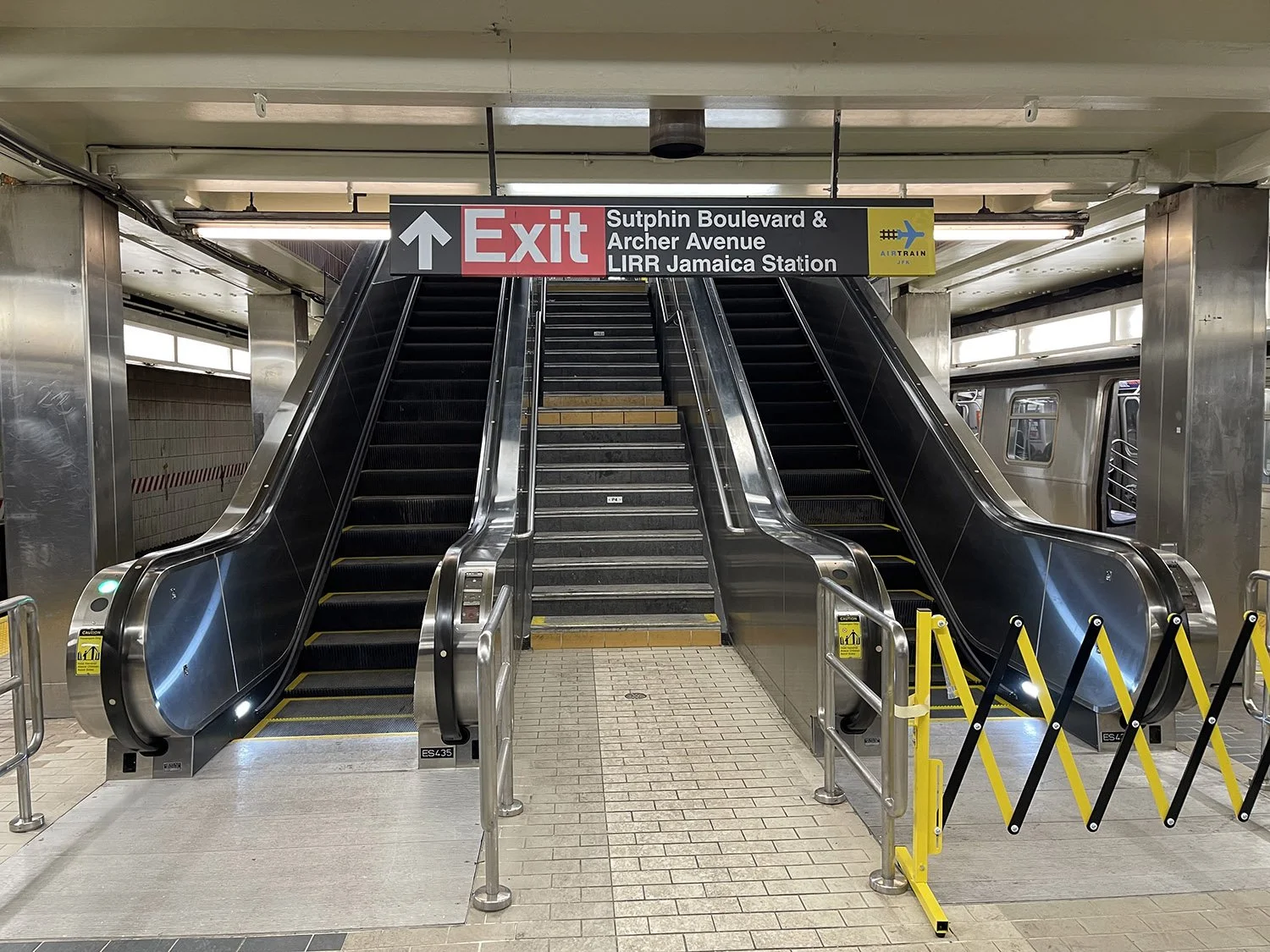 Escalators in a subway station leading to the exit, with a sign overhead indicating directions to Sutphin Boulevard, Archer Avenue, and Jamaica Station, as well as a yellow barrier on the right.