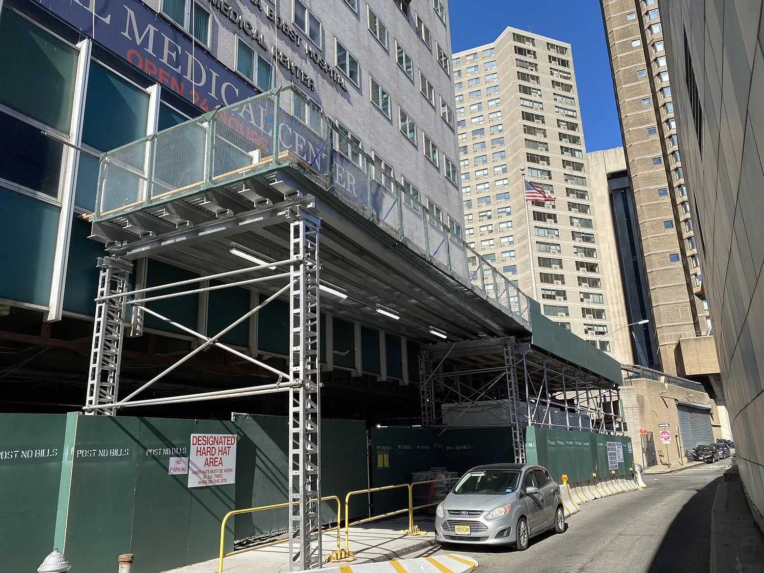 Construction site in front of tall city buildings with a sign that reads 'Designated Hard Hat Area'.
