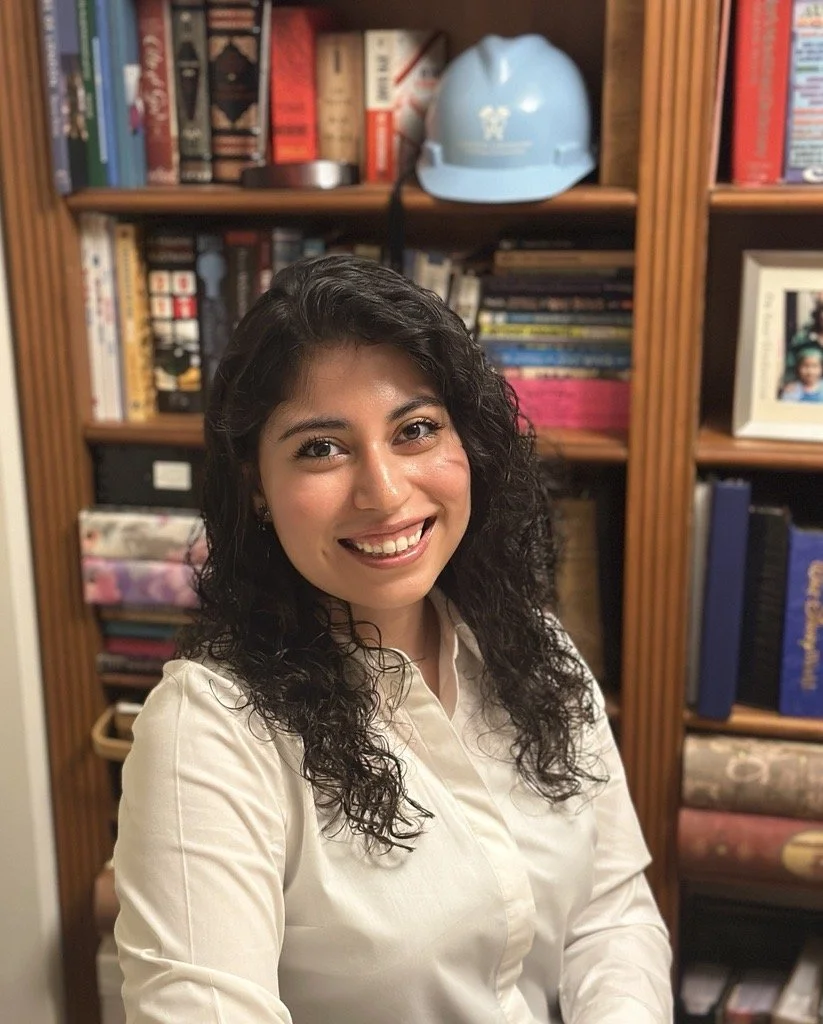 A young woman smiling at the camera, with dark curly hair, wearing a cream-colored shirt, standing in front of a wooden bookshelf filled with books and a blue safety helmet on the top shelf.