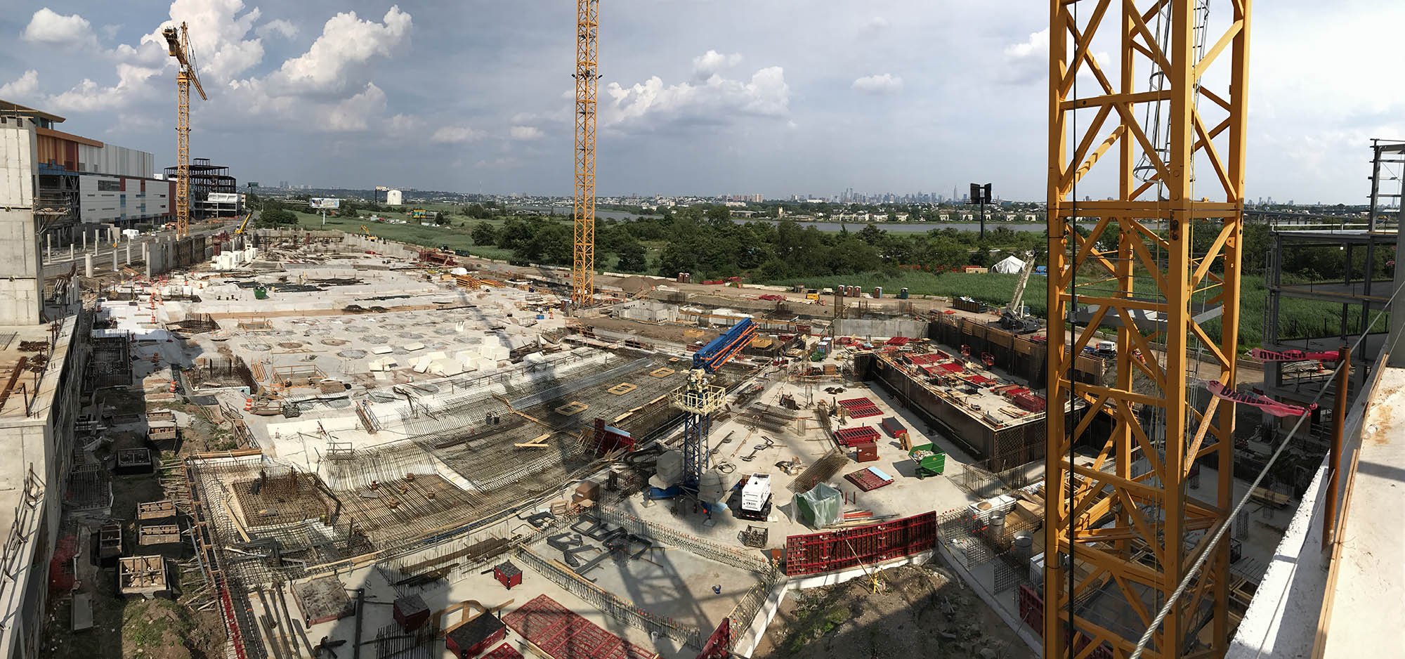 A large construction site with cranes and building materials, with a city skyline and river in the background under a partly cloudy sky.