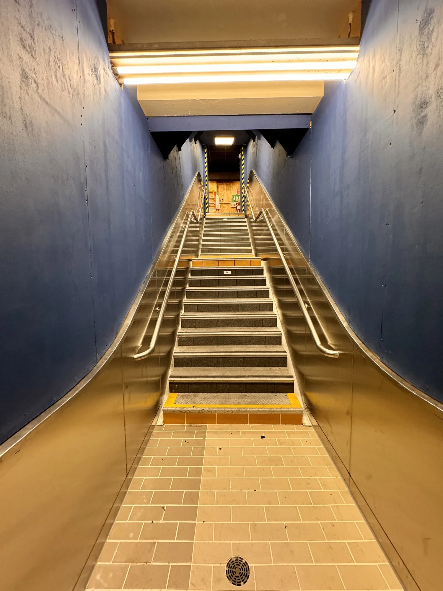 Staircase with handrails leading up to an underground area with blue walls and fluorescent lighting.
