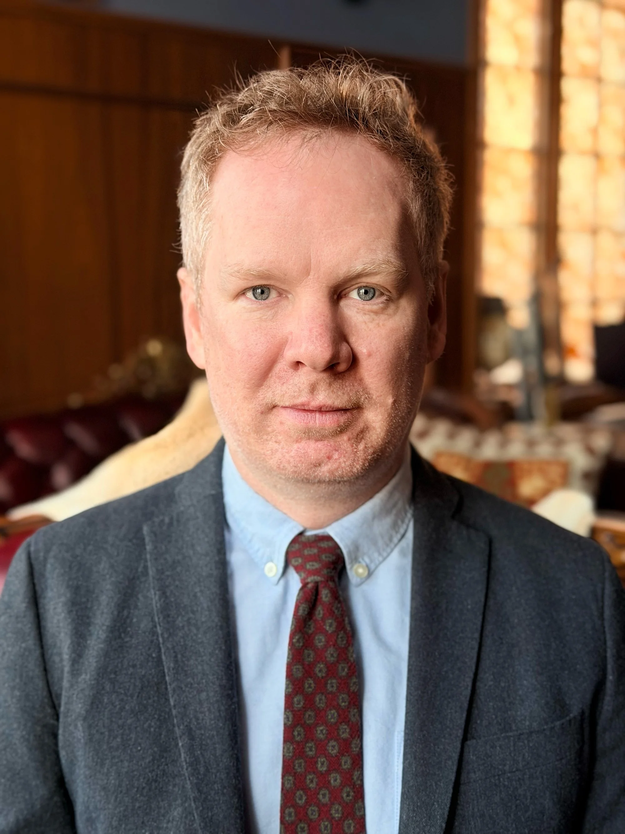 Portrait of a man with reddish hair and blue eyes, wearing a dark blazer, light blue shirt, and patterned red tie, set against a warmly lit background with wooden paneling and windows.