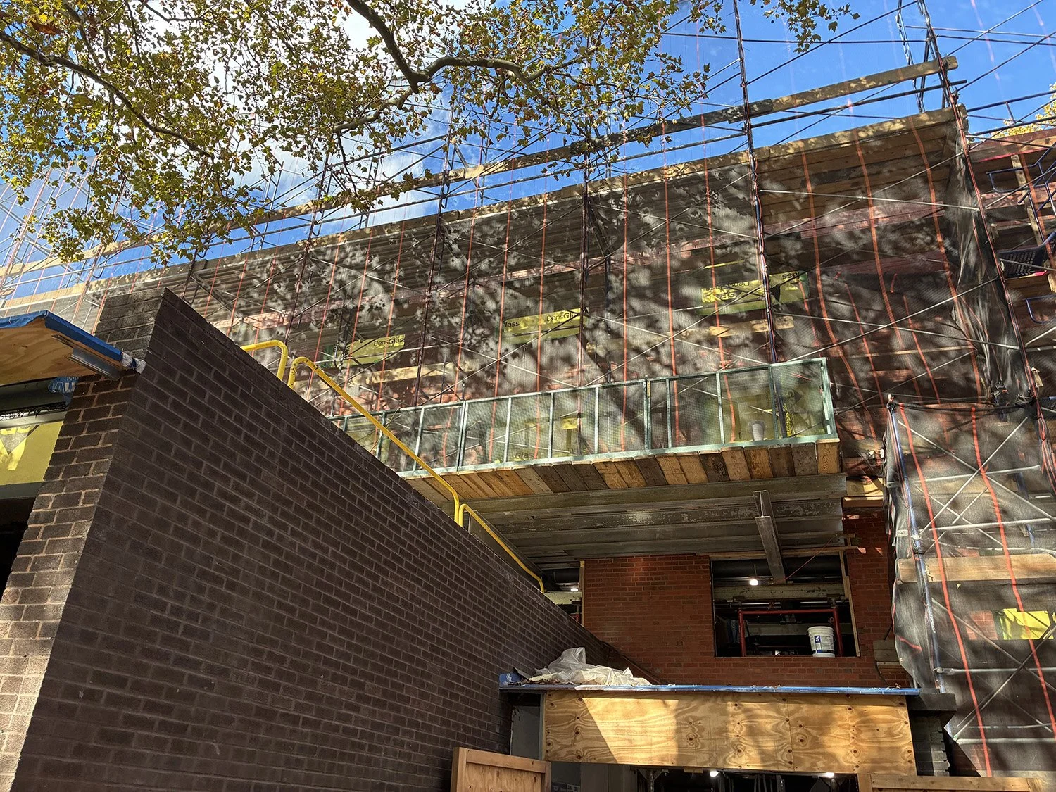 Building under construction with scaffolding around it, brick walls, and a blue sky with some clouds and tree branches visible.