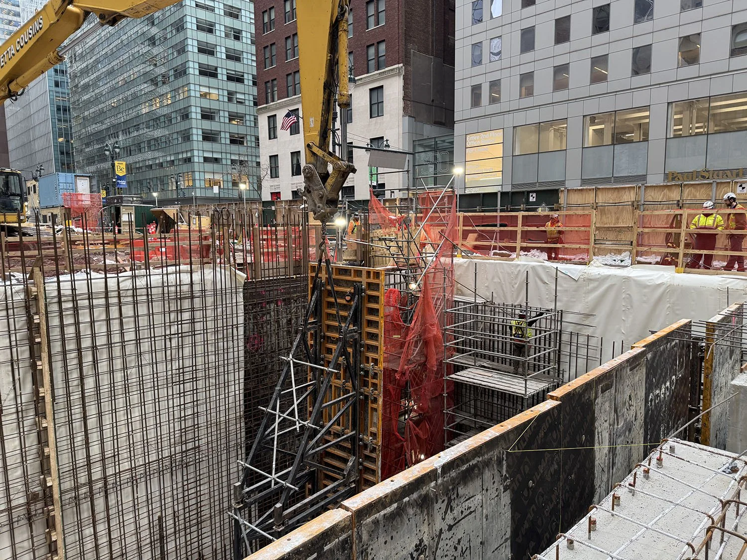 Construction workers wearing yellow vests and hard hats working on a building site surrounded by metal rebar, wooden scaffolding, and construction equipment in an urban area with tall office buildings.
