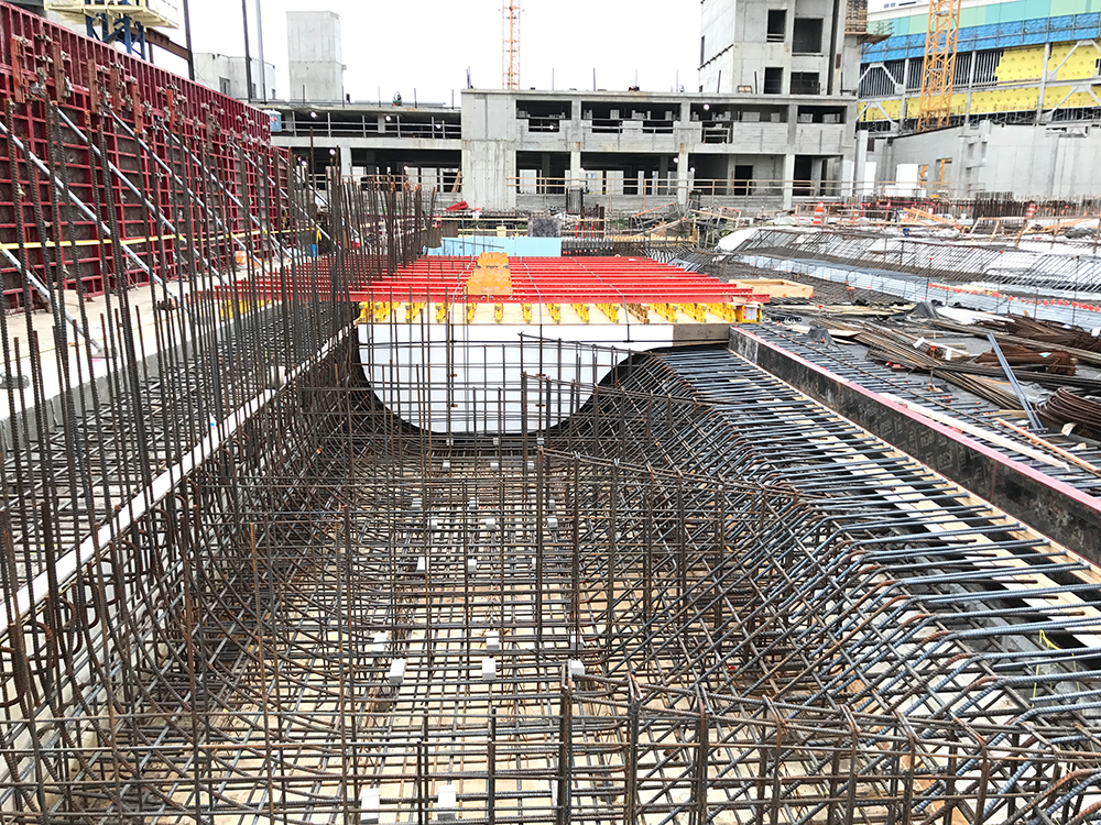 Construction site with steel rebar framework for concrete foundation, surrounded by partially built structures and construction materials.