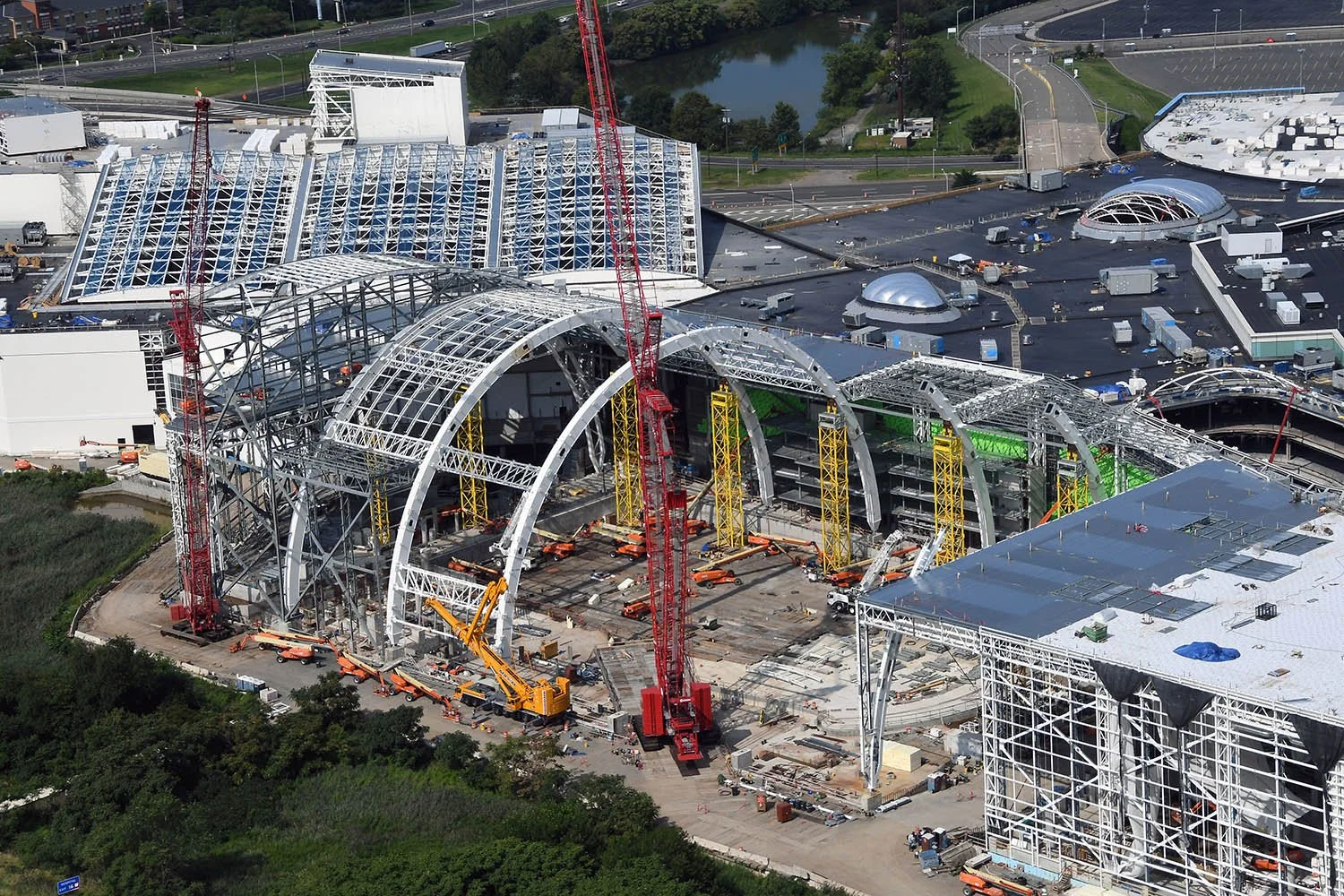 Aerial view of a large construction site with steel framework, cranes, and building structures in progress, surrounded by roads, parking lot, and greenery.