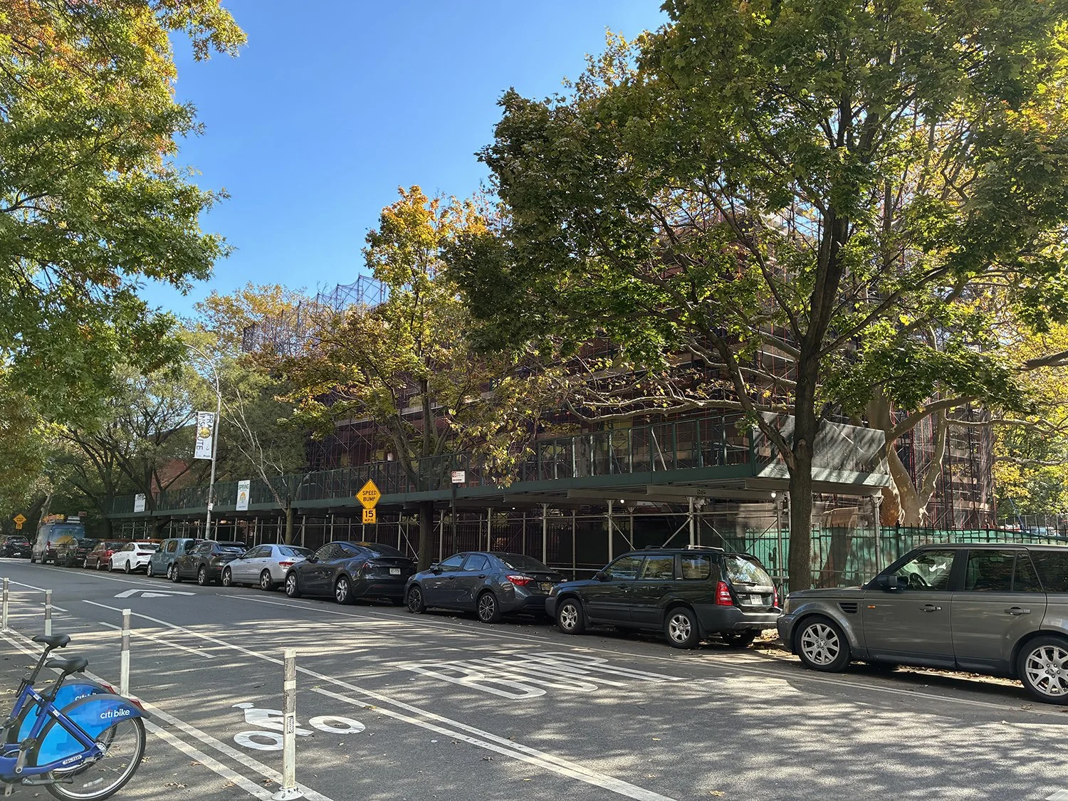 City street with parked cars along a sidewalk, trees with green leaves, and a building under construction surrounded by scaffolding. Clear blue sky above.