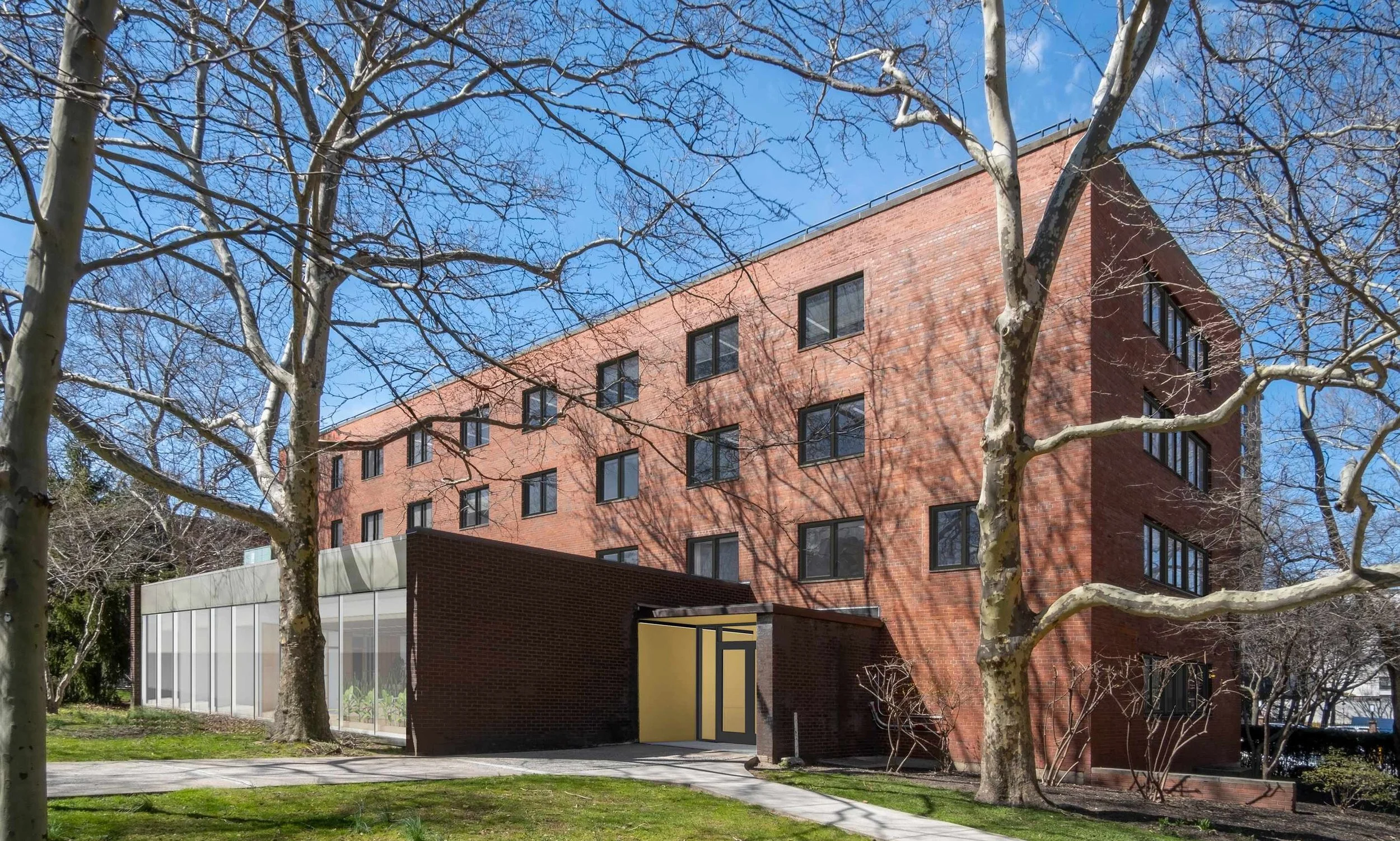 A modern multi-story brick building with black window frames, surrounded by leafless trees, under a bright blue sky.