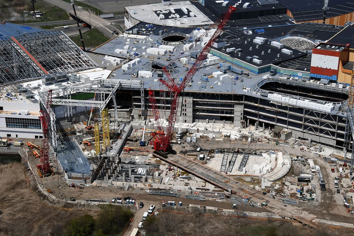 Aerial view of a large building under construction, with cranes, construction equipment, and workers, surrounded by roads and greenery.