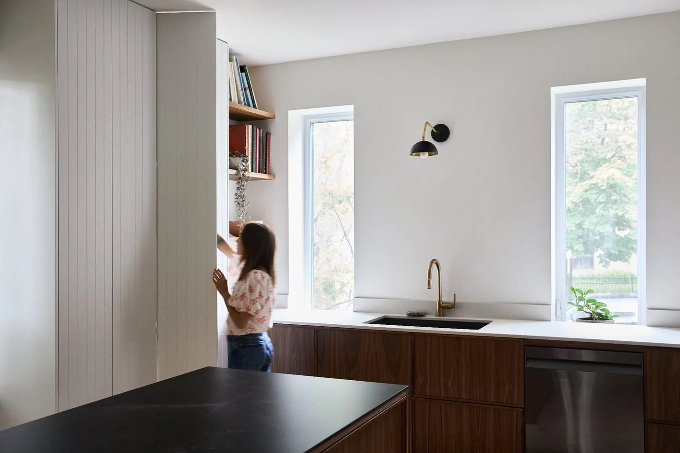 Une femme avec une fille dans une cuisine moderne avec deux fenêtres, évier en or et étagères avec des livres