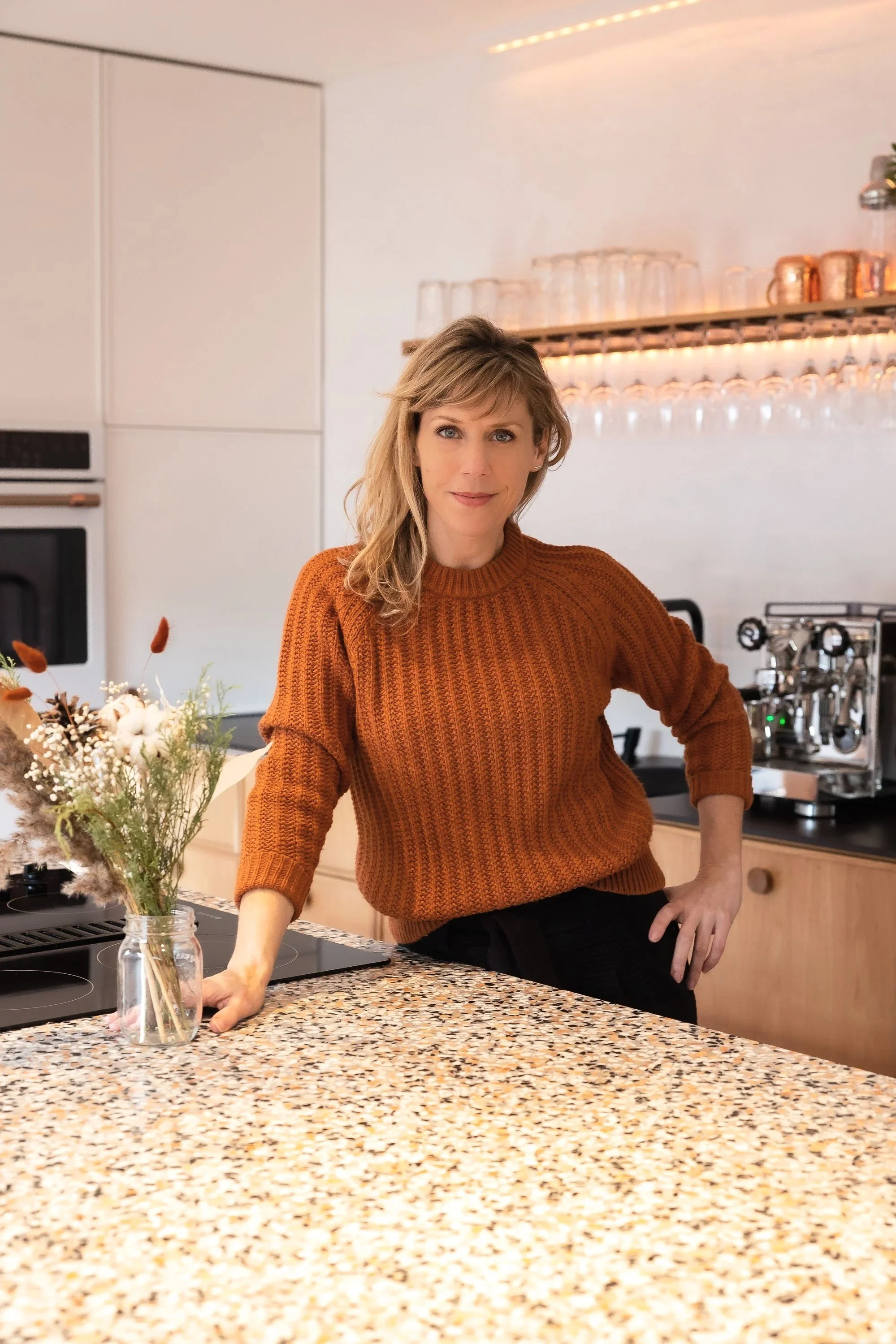 Une femme se tient dans une cuisine moderne portant un pull orange, avec un peu de maquillage, souriant légèrement, devant un plan de travail en pierre, avec un bouquet de fleurs dans un vase en verre à portée de main.