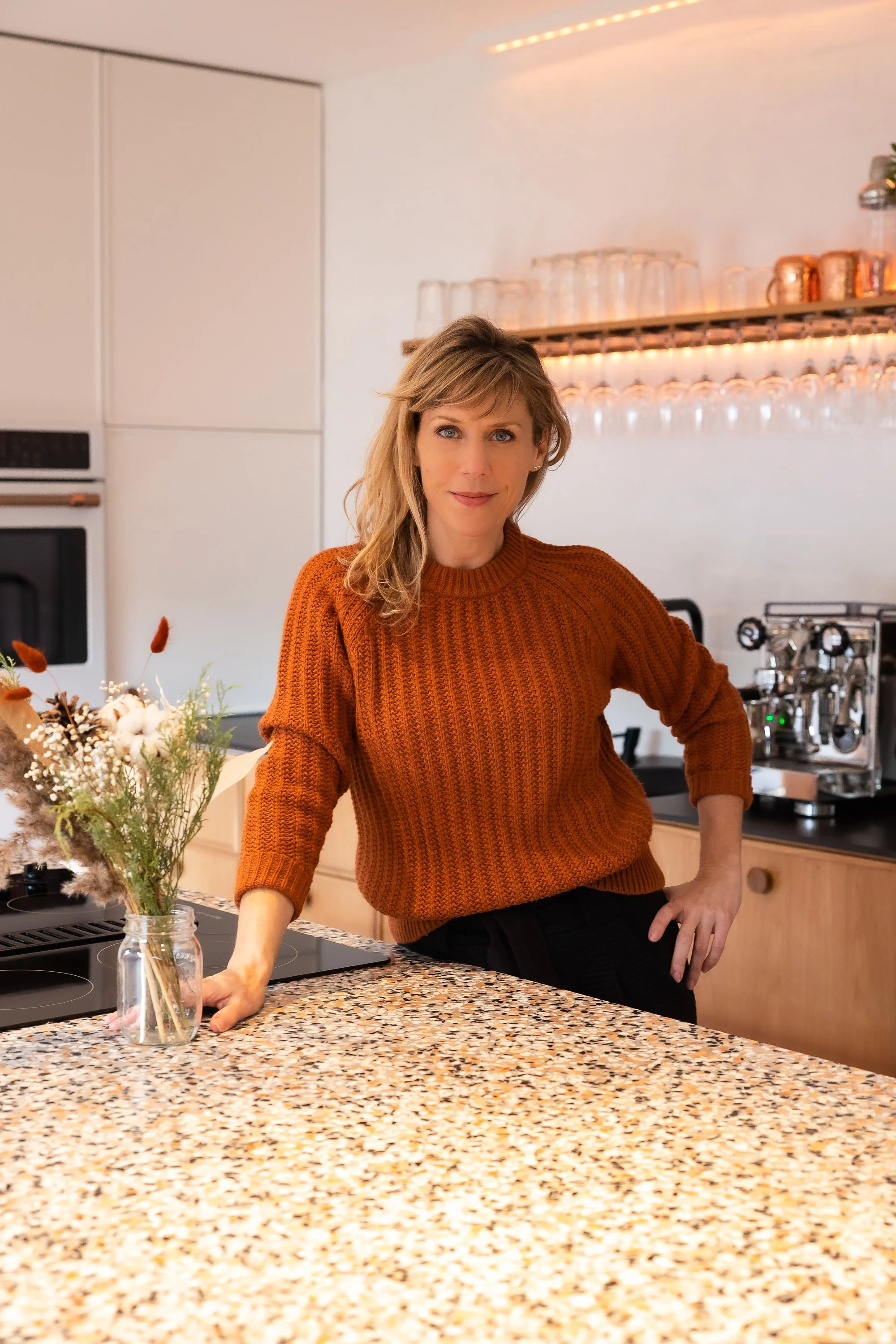 Une femme souriante en pull orange debout dans une cuisine moderne, posant à côté d'un vase avec des fleurs, avec des verres en arrière-plan sur une étagère en bois.