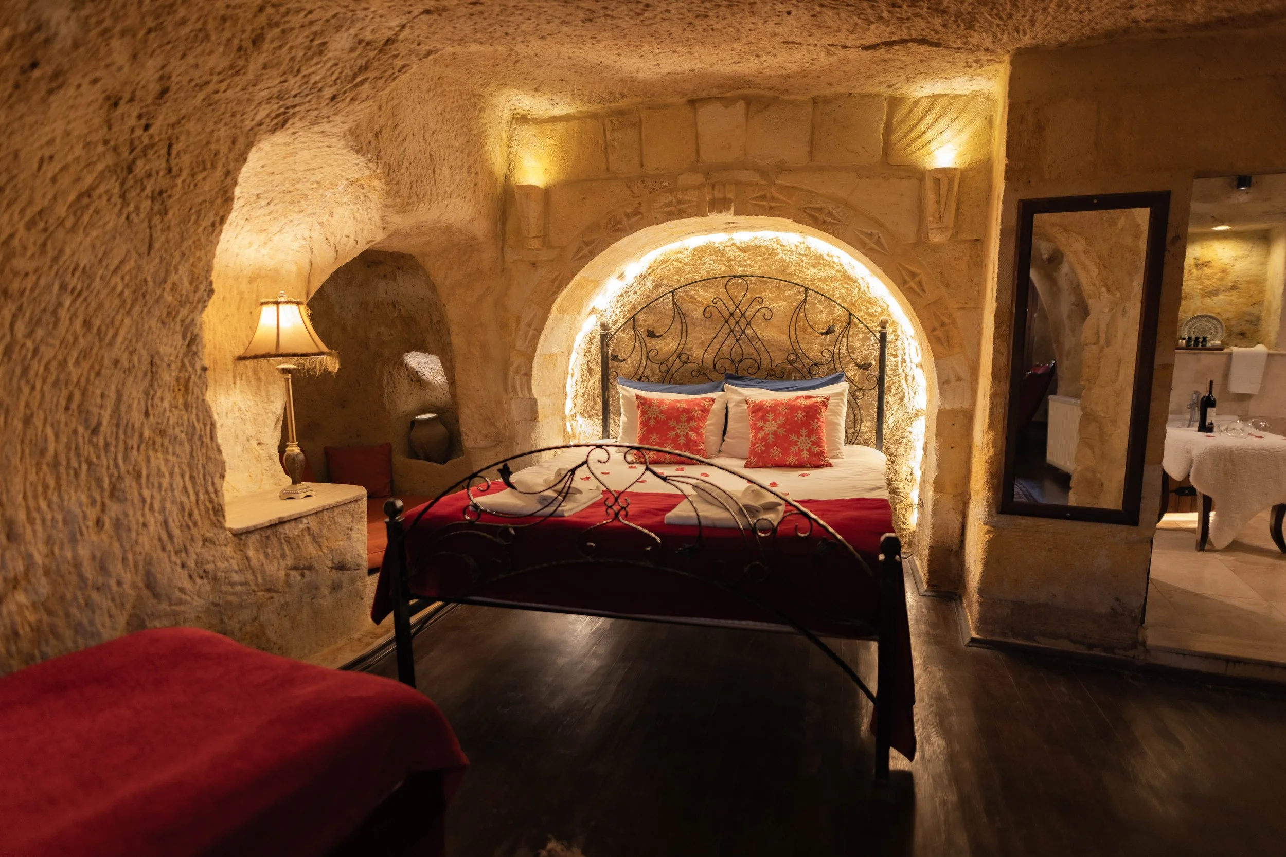 Ein gemütliches Schlafzimmer mit einer Steinwand, einem Bett mit roten und weißen Bettdecken und dekorativen Kissen, seitlich eine Nachttischlampe, im Hintergrund eine Spiegelwand und ein Essbereich.