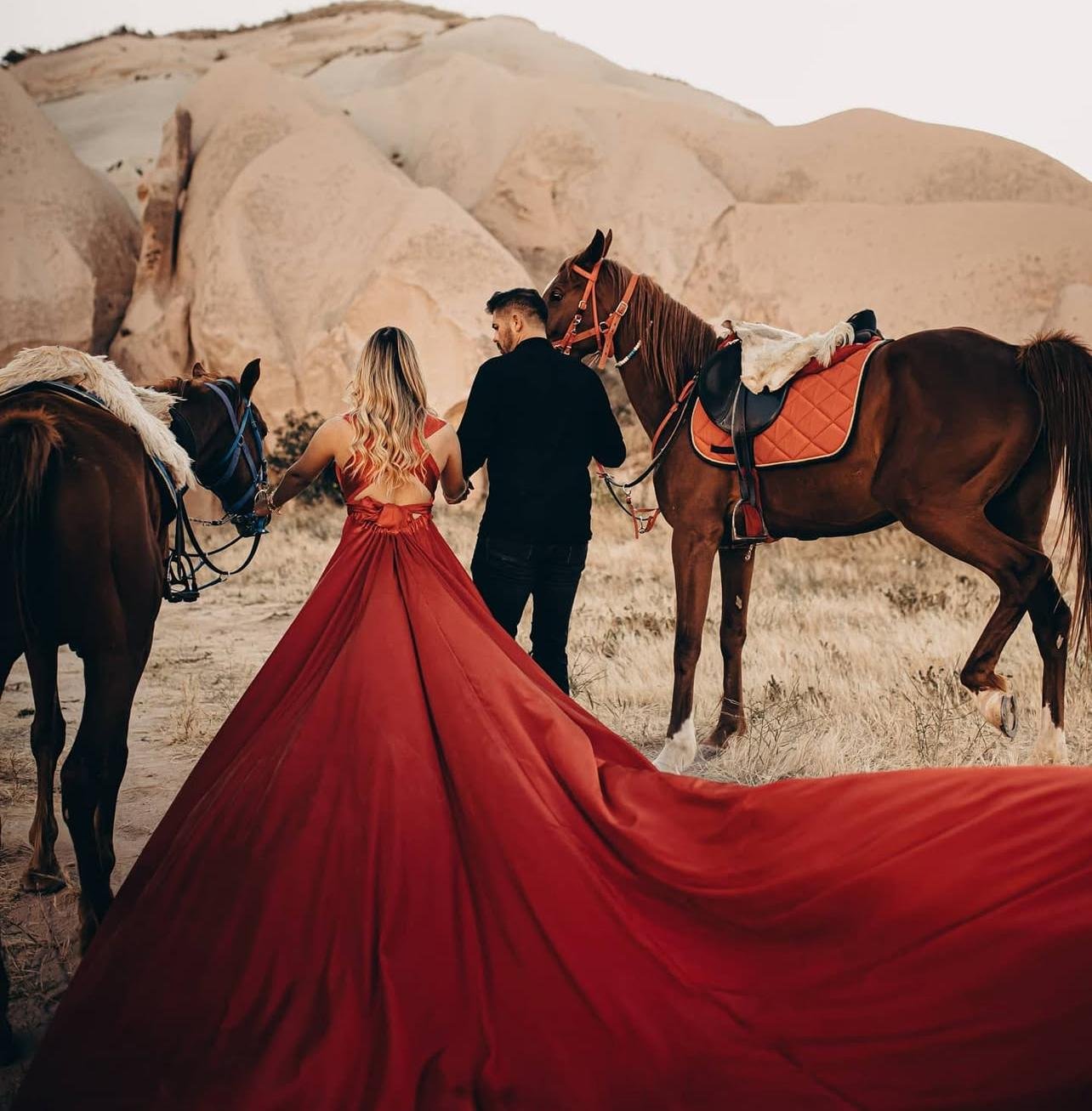Ein Mann und eine Frau stehen mit ihren Pferden in einer Wüstenlandschaft, die Frau trägt ein langes rotes Kleid.