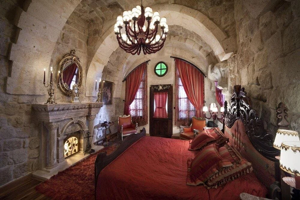 Ein rustikales Schlafzimmer mit Steinwänden, einem großen Holz-Kopfkissen, roten Bettwäsche und Vorhängen, einem Kamin, Spiegel, Kaminschmuck und antiken Möbeln.