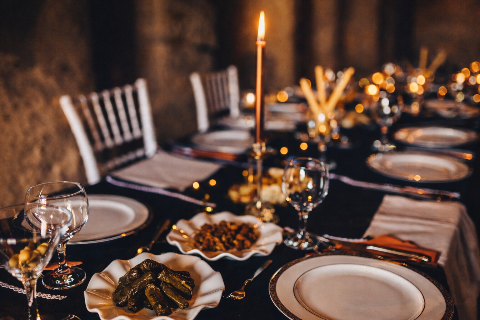 Ein festlich gedeckter Tisch mit weißen Tellern, Gläsern und Silberbesteck, beleuchtet von Kerzen, mit Speisen wie gefüllten Weinblättern und einer vegetarischen Fleischplatte, in einer rustikalen Umgebung.