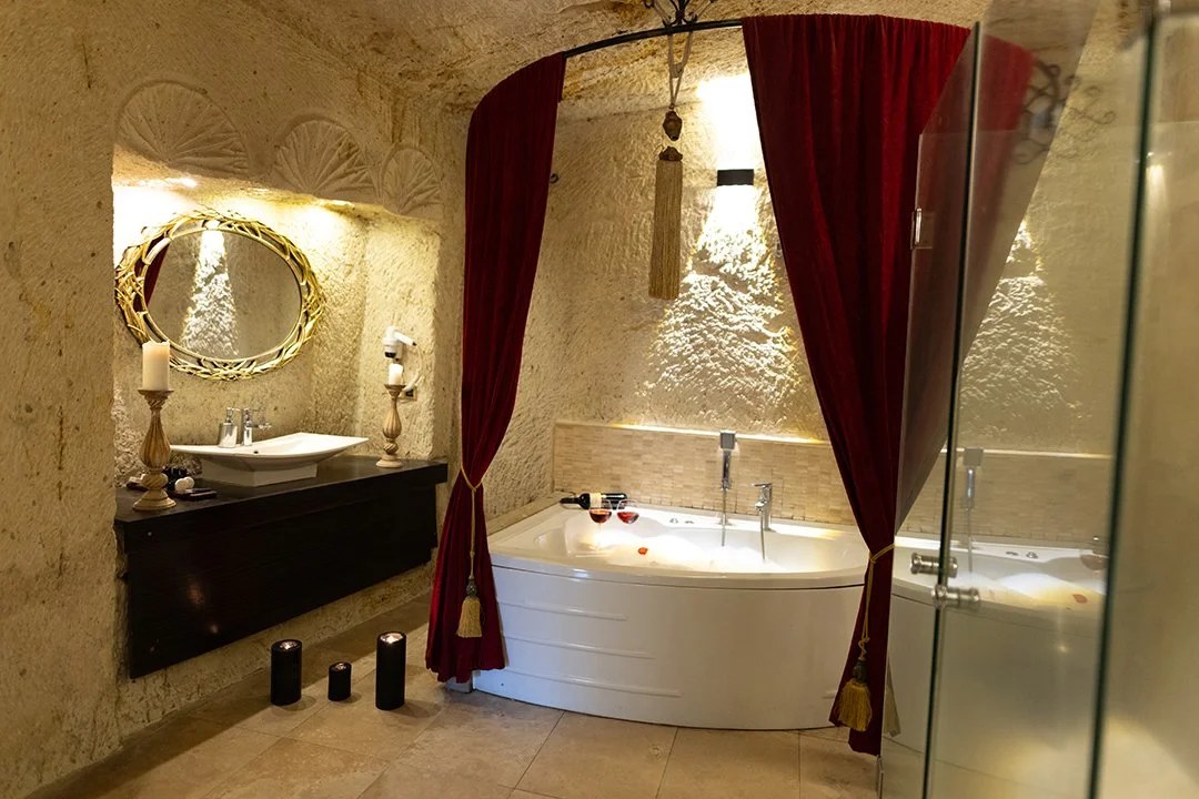 Ein Badezimmer mit einer Badewanne, die durch rote Vorhänge abgetrennt ist, einem Waschtisch mit Spiegel und Kerzen, sowie Decken- und Wandbeleuchtung in warmem Licht.