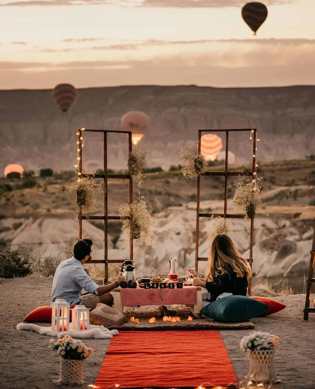 Paar beim Picknick im Freien bei Sonnenuntergang, mit Heißluftballons im Himmel und dekoriertem Tisch.