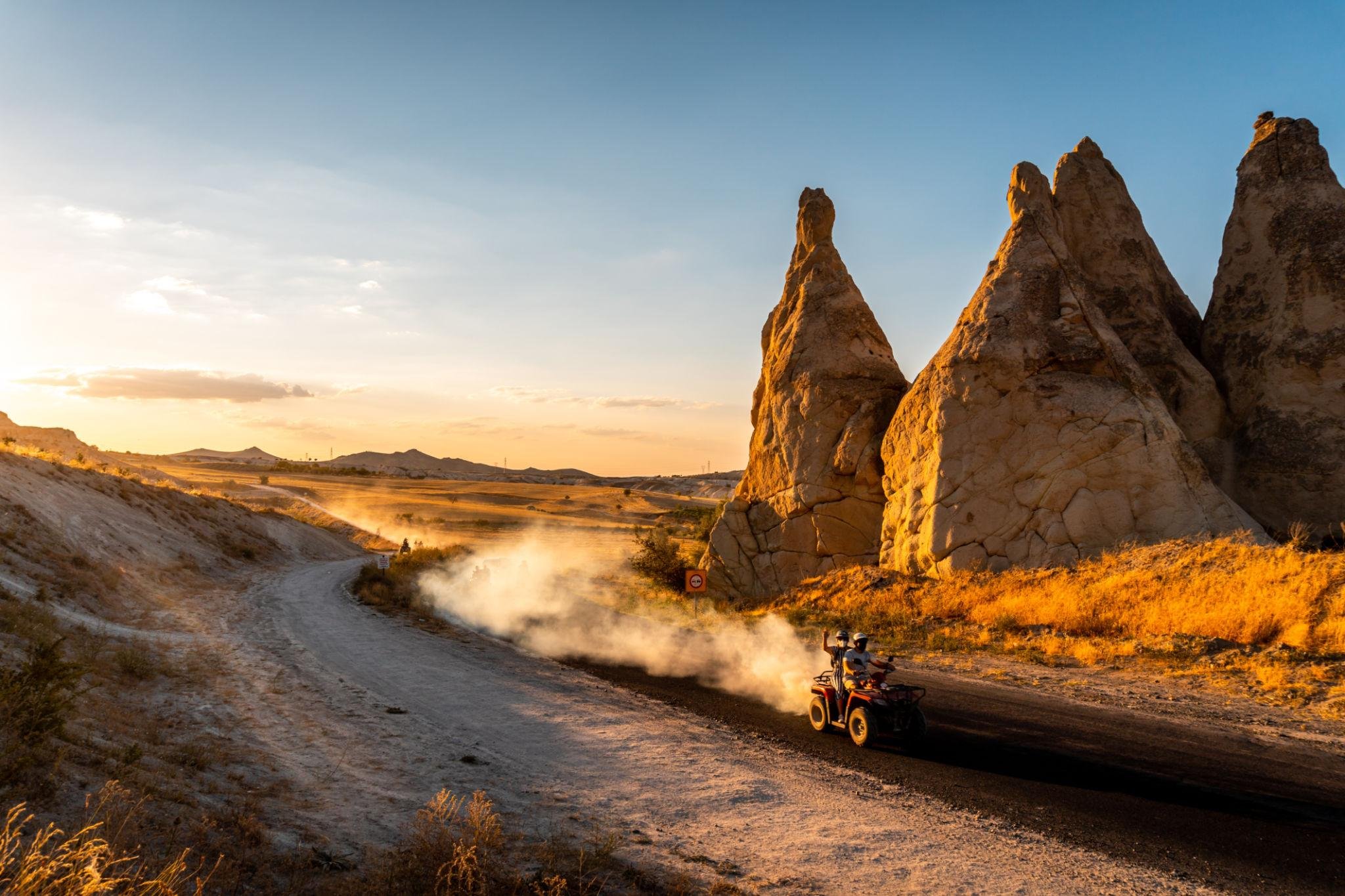 Zwei Personen fahren mit einem ATV auf einer staubigen Straße durch eine wüstenartige Landschaft mit großen Felsen bei Sonnenuntergang.