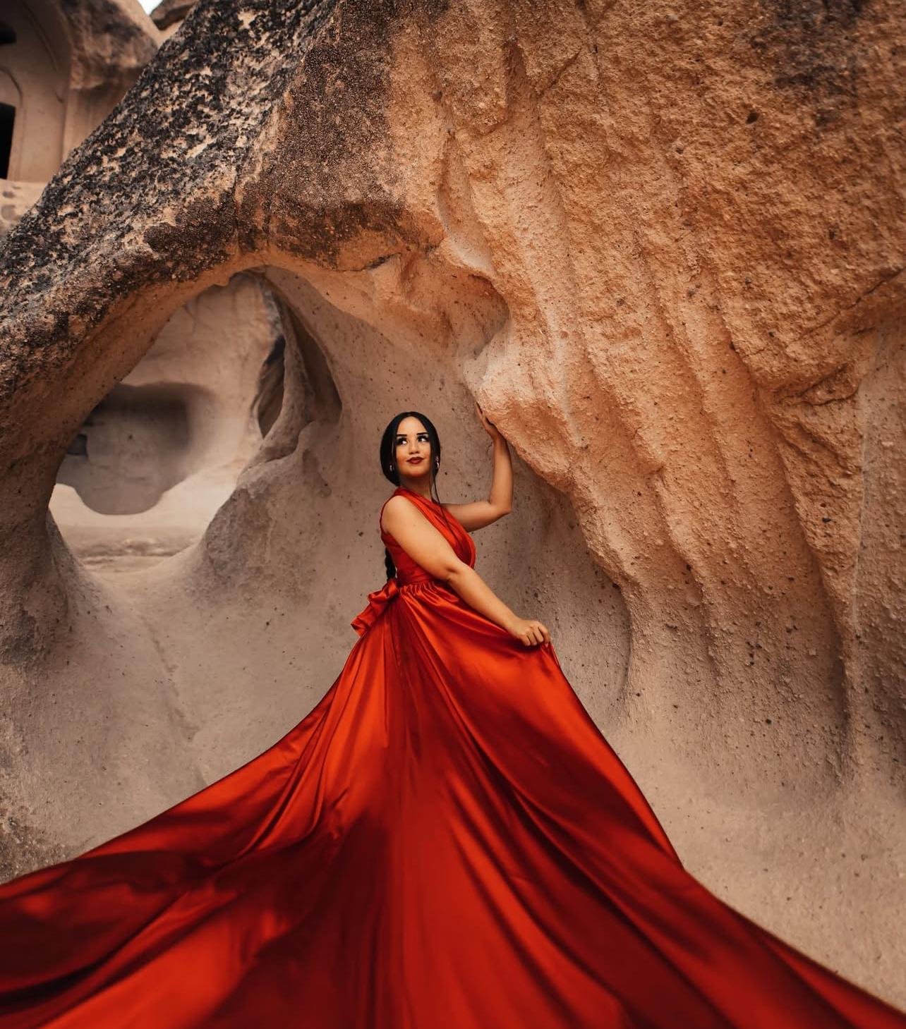 Eine Frau in einem langen, roten Kleid steht in einer natürlichen Felsenformation. Sie berührt eine Felswand mit ihrer Hand und blickt nach oben.