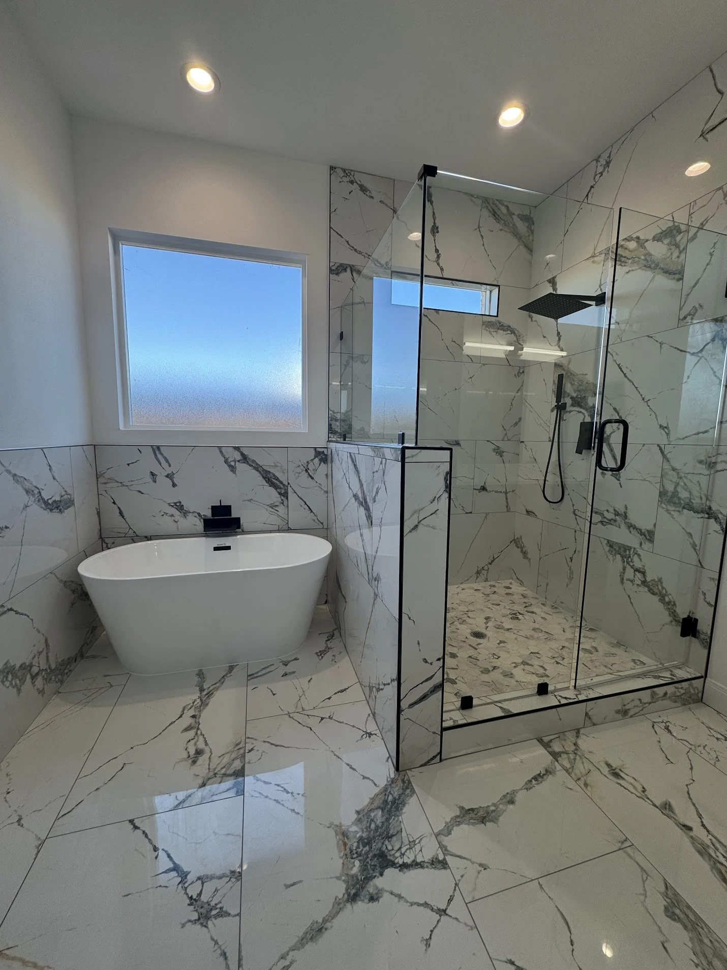 Modern bathroom with marble tile walls and floor, a freestanding bathtub below a frosted window, and a glass-enclosed shower with rainfall showerhead.